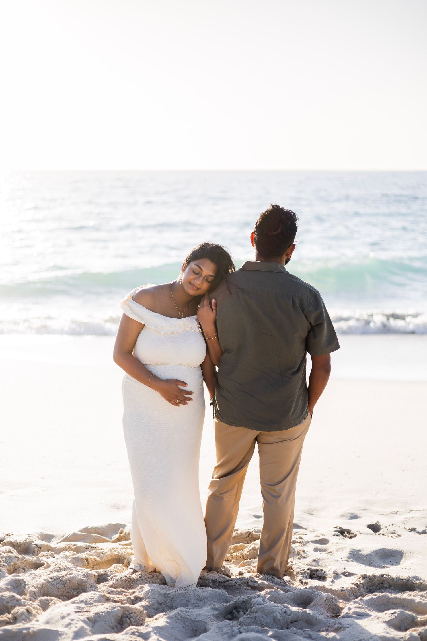 Pregnant woman in white dress leans on partner's shoulder while standing on sandy beach at sunset.