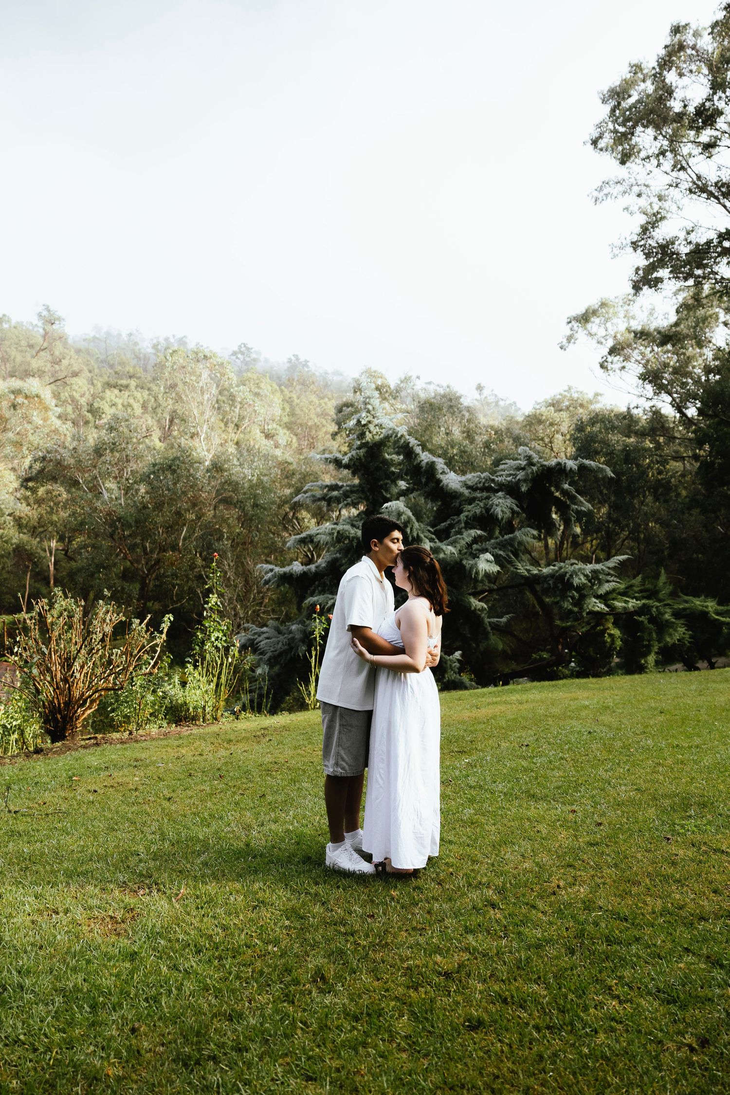 Couple embracing on green lawn surrounded by lush trees and garden, romantic outdoor portrait.