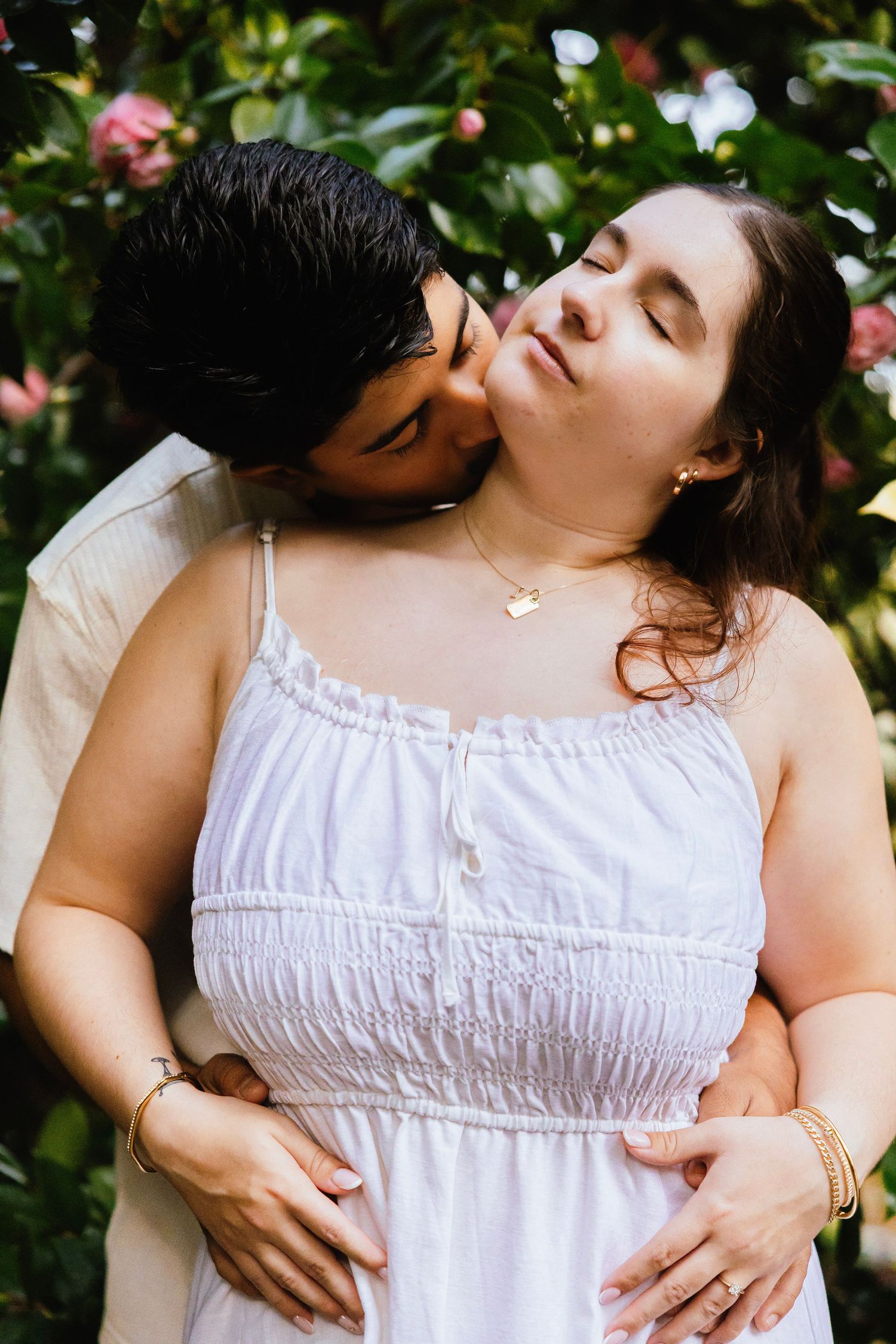 A couple shares a tender moment outdoors, man kissing pregnant woman's cheek near pink flowering bushes.