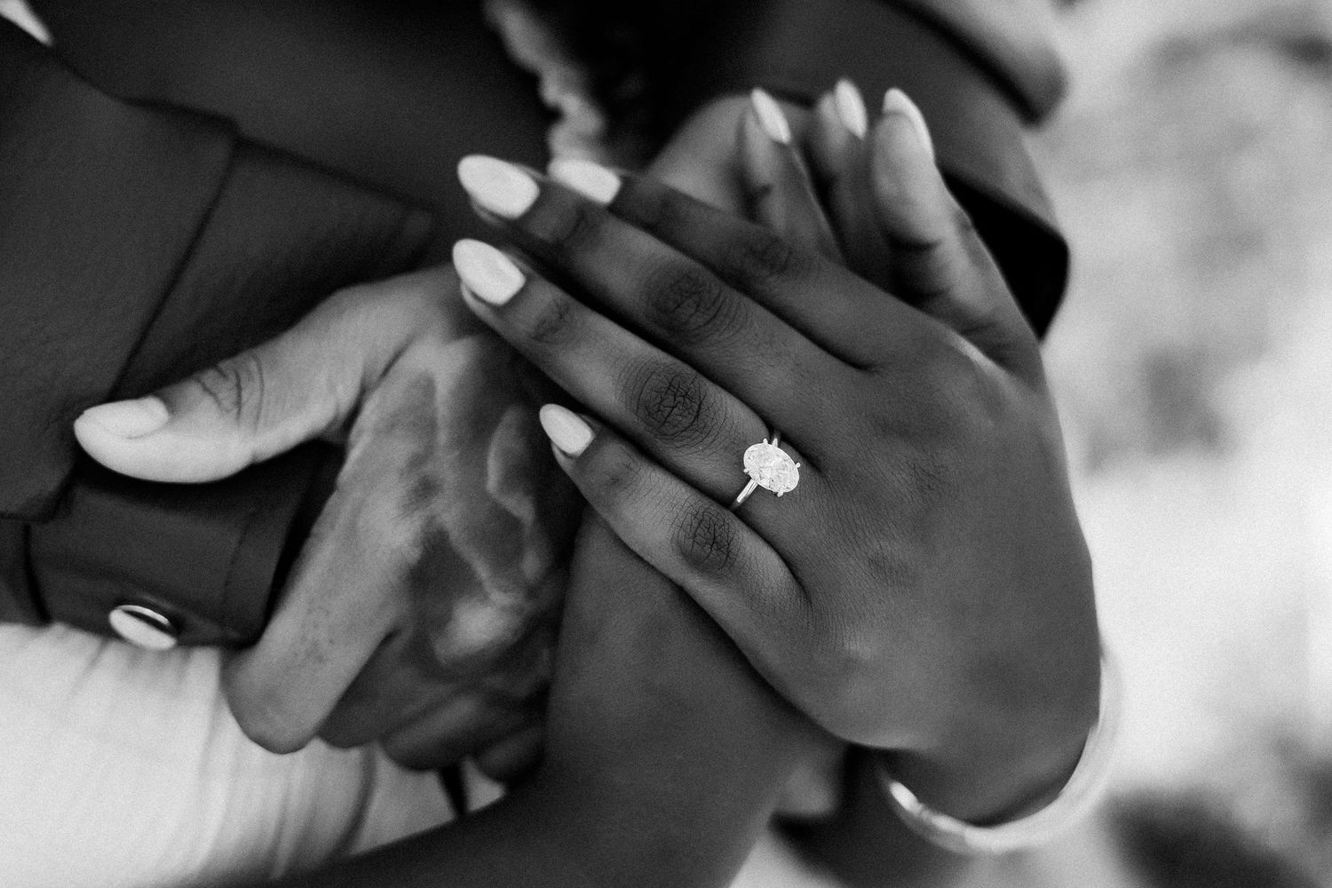 Black and white photo of a woman showing off a diamond engagement ring on her finger while holding her partner's arm.