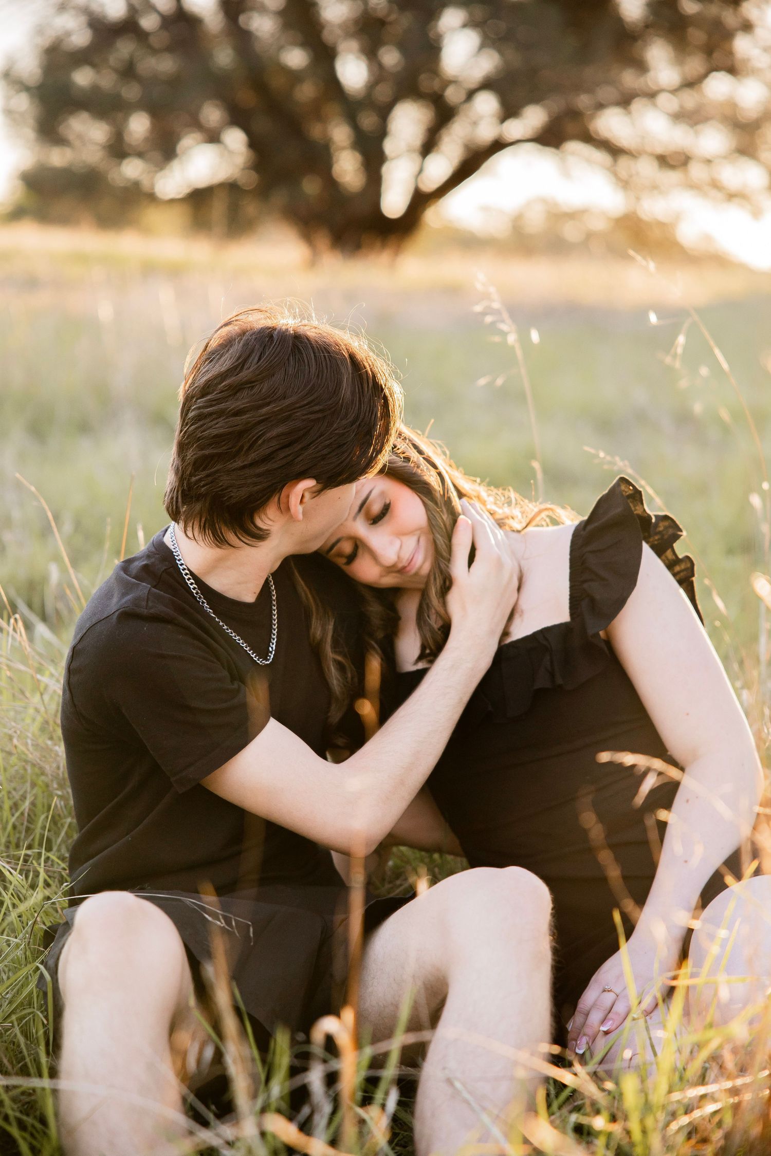 Couple sitting in golden field at sunset, embracing tenderly in matching black outfits with warm backlight.