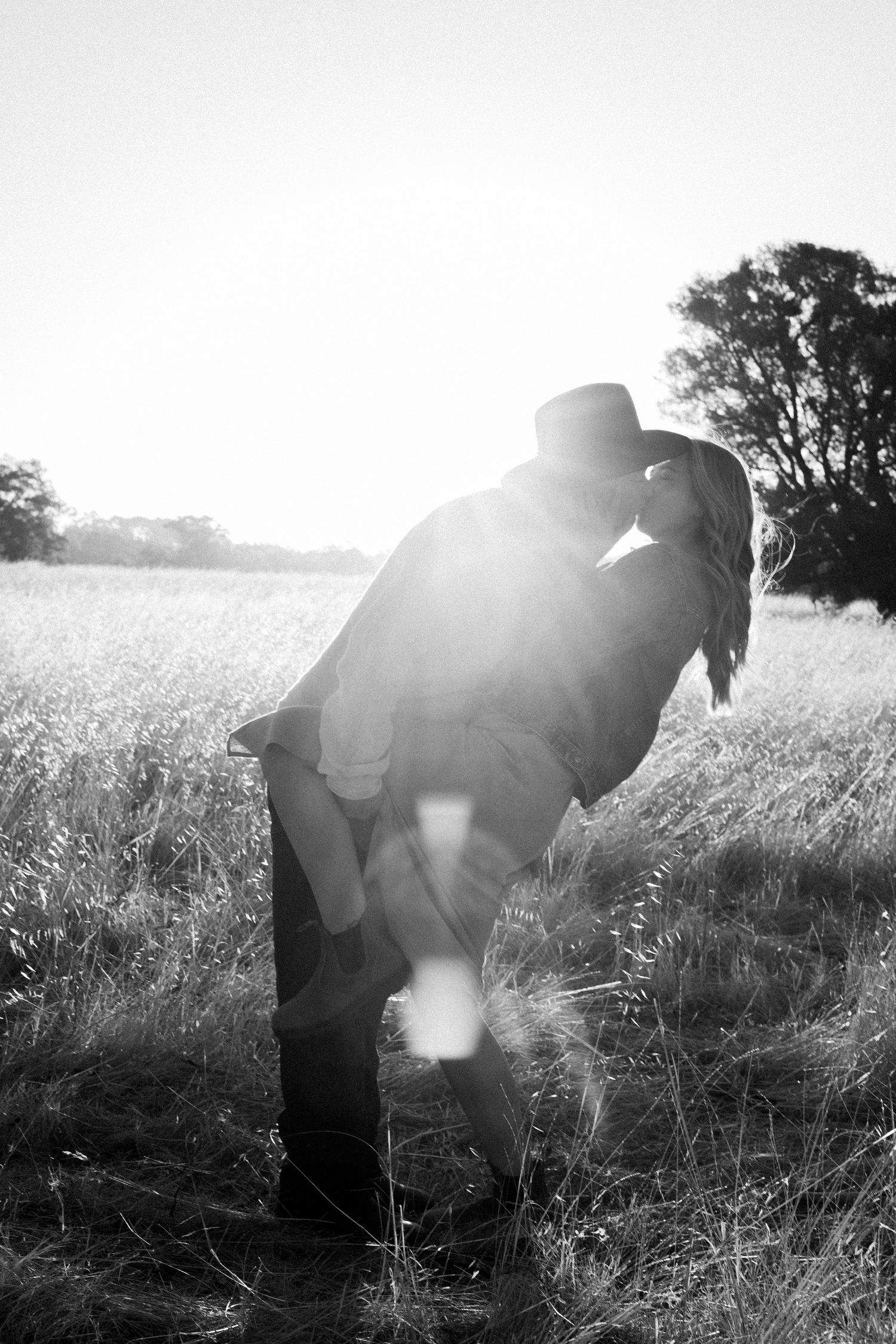 Black and white image of a couple kissing in a sunlit field with dramatic lens flare.