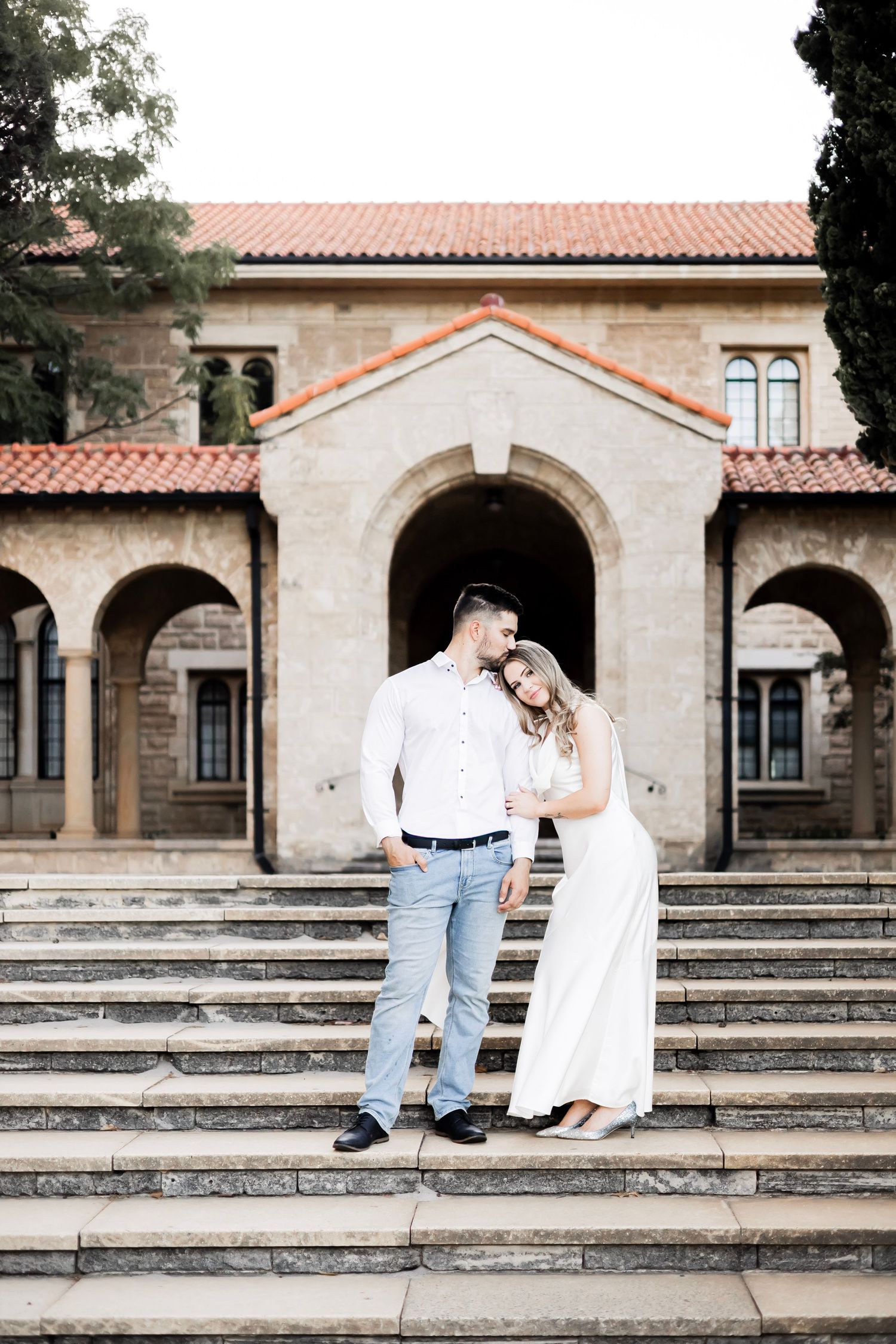 Couple embracing on stone steps before a Spanish-style building with arched entryways and red tile roof.