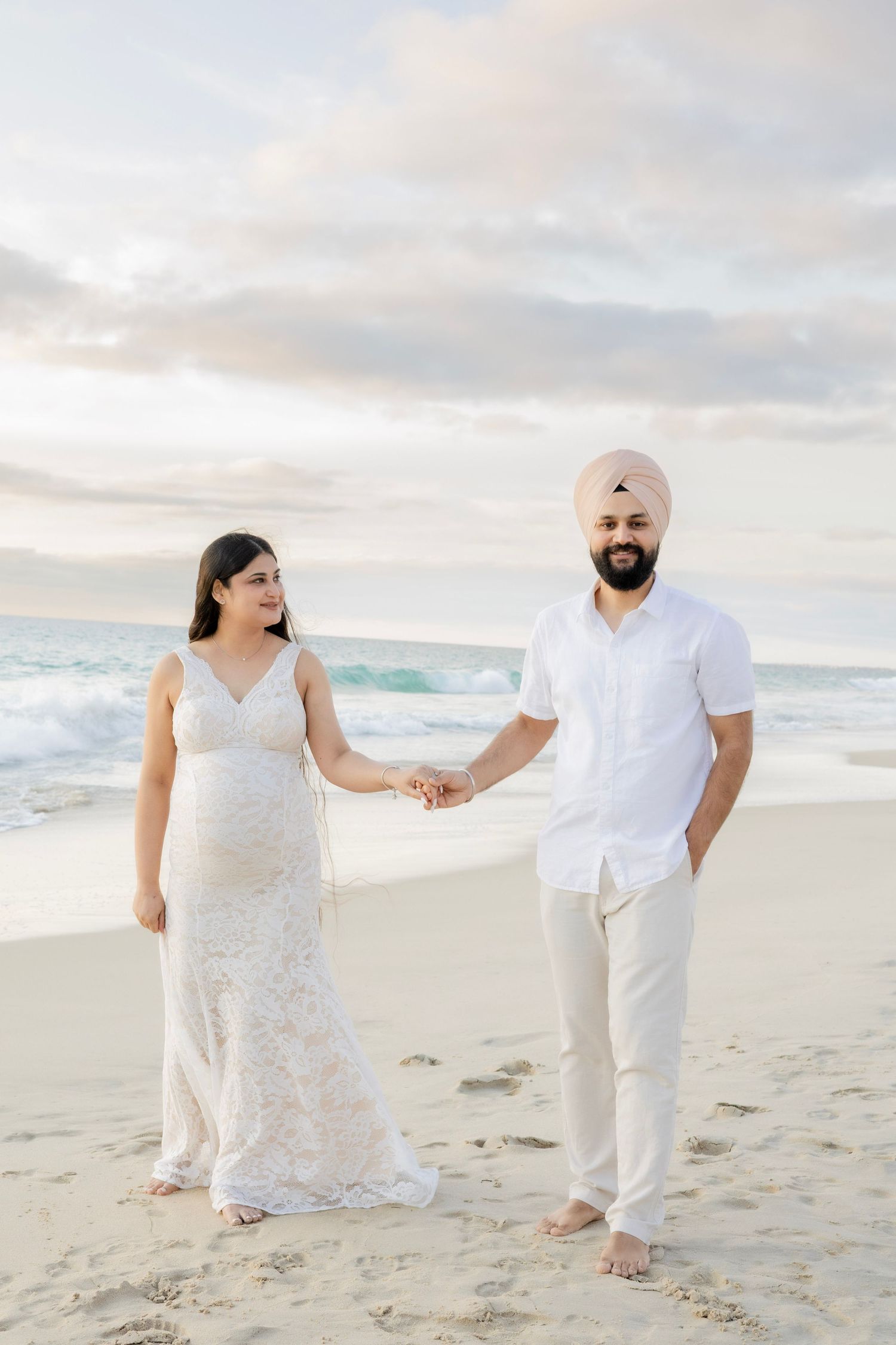 Expecting couple holding hands on sandy beach at sunset, man in turban and white outfit, woman in lace dress.