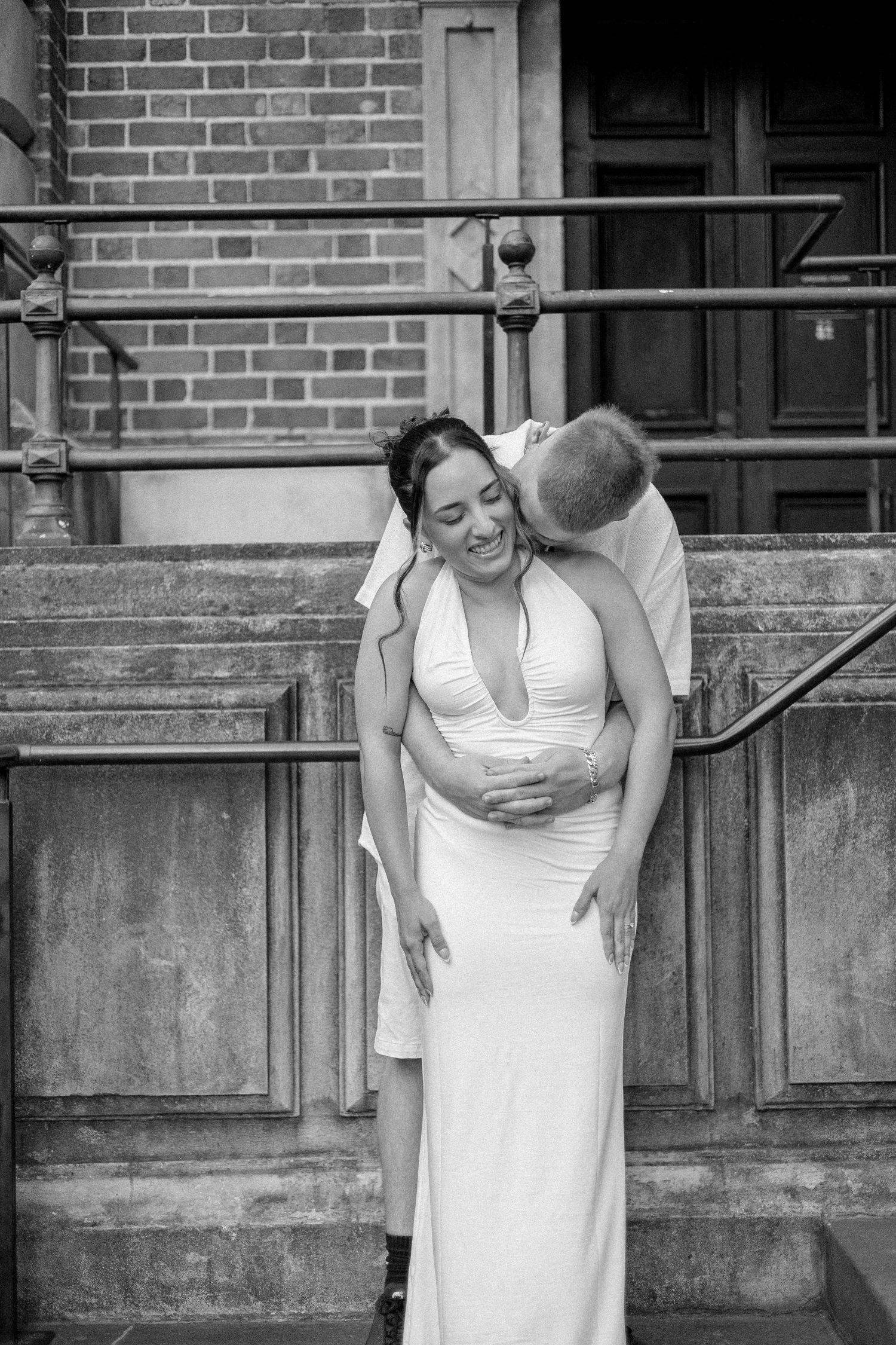Black and white photo of man embracing laughing pregnant woman in white halter dress on stone steps.