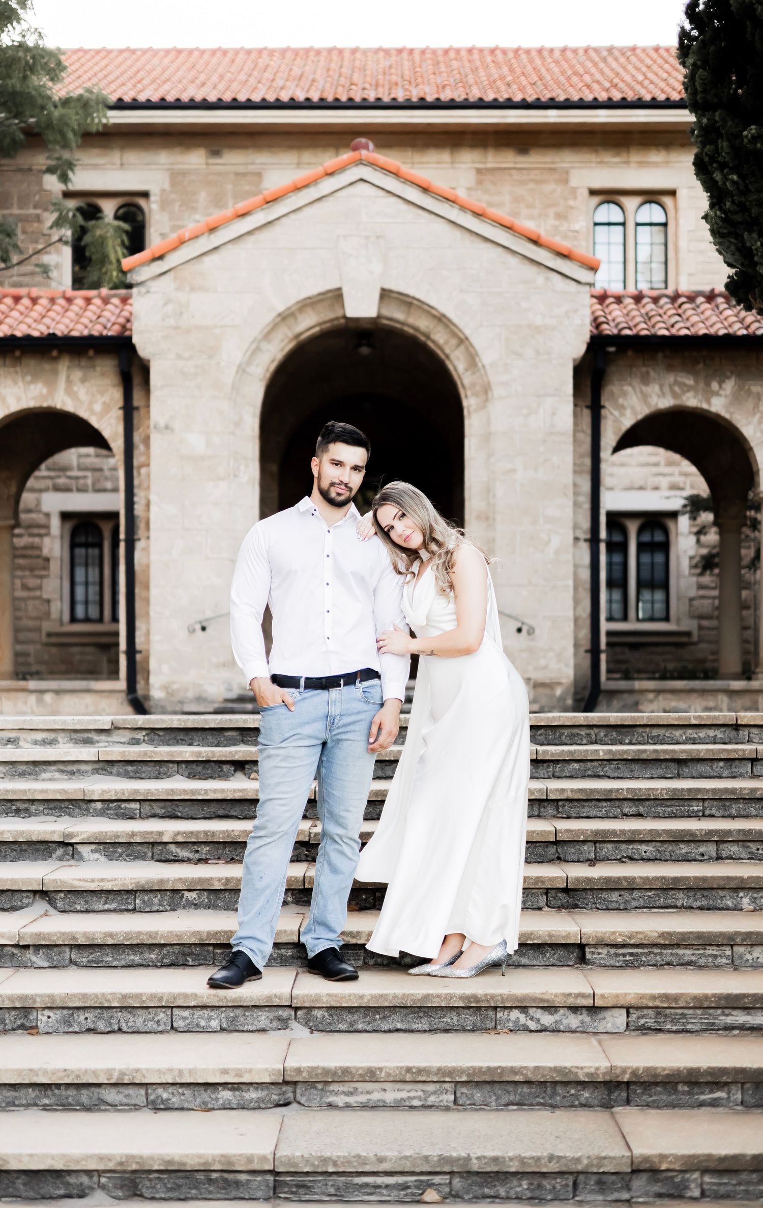 Couple in matching white outfits posing on stone steps in front of a historic building.