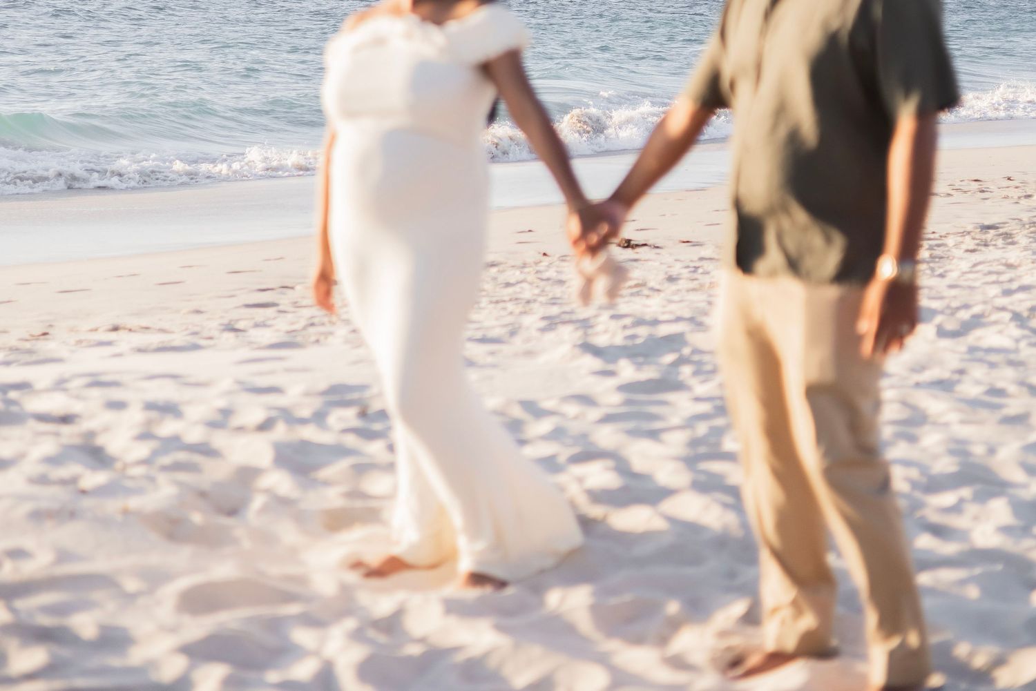 Pregnant woman in white dress holds hands with partner on sandy beach with ocean waves in background.