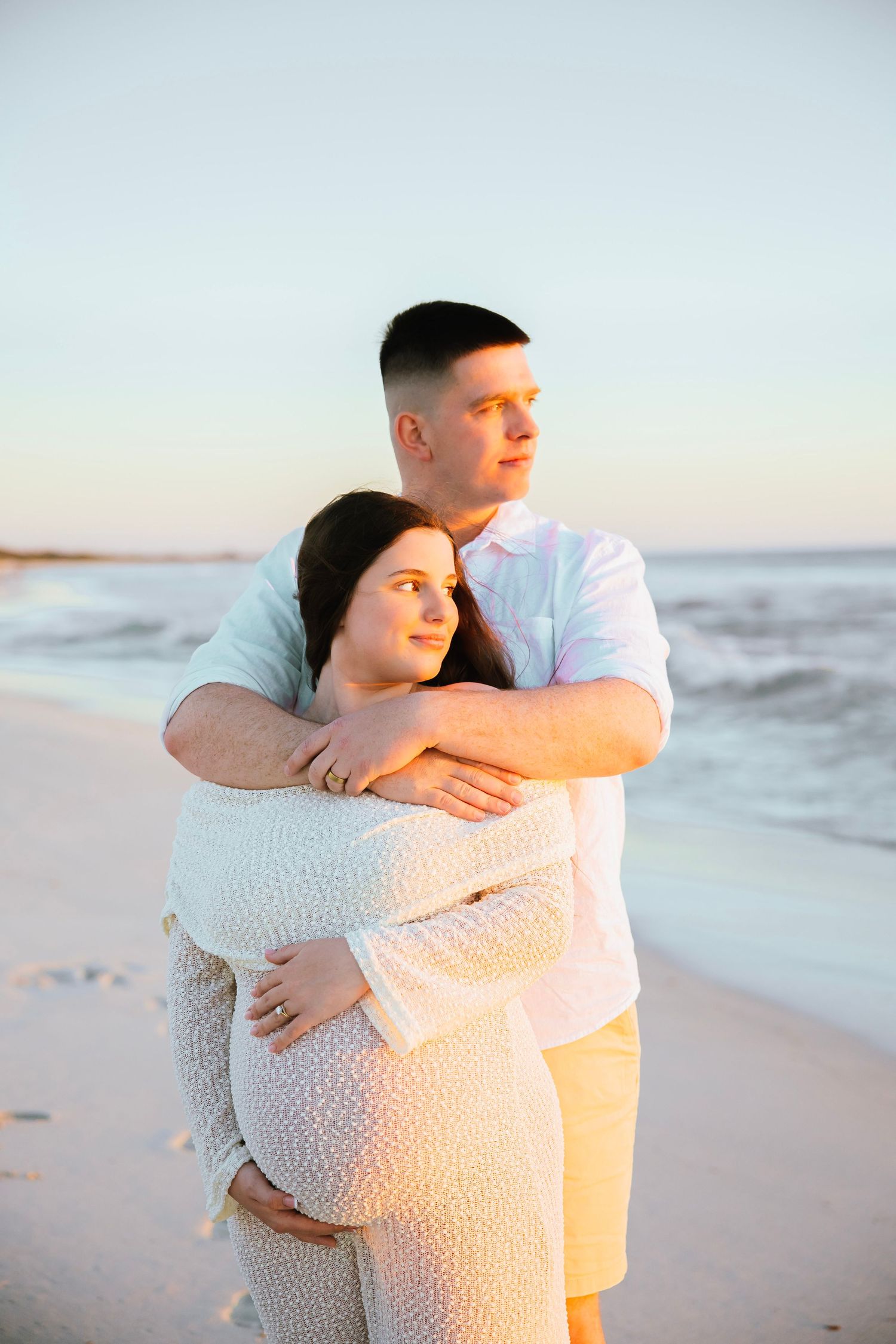Couple embracing on beach at sunset, pregnant woman in white lace dress held by man in white shirt.