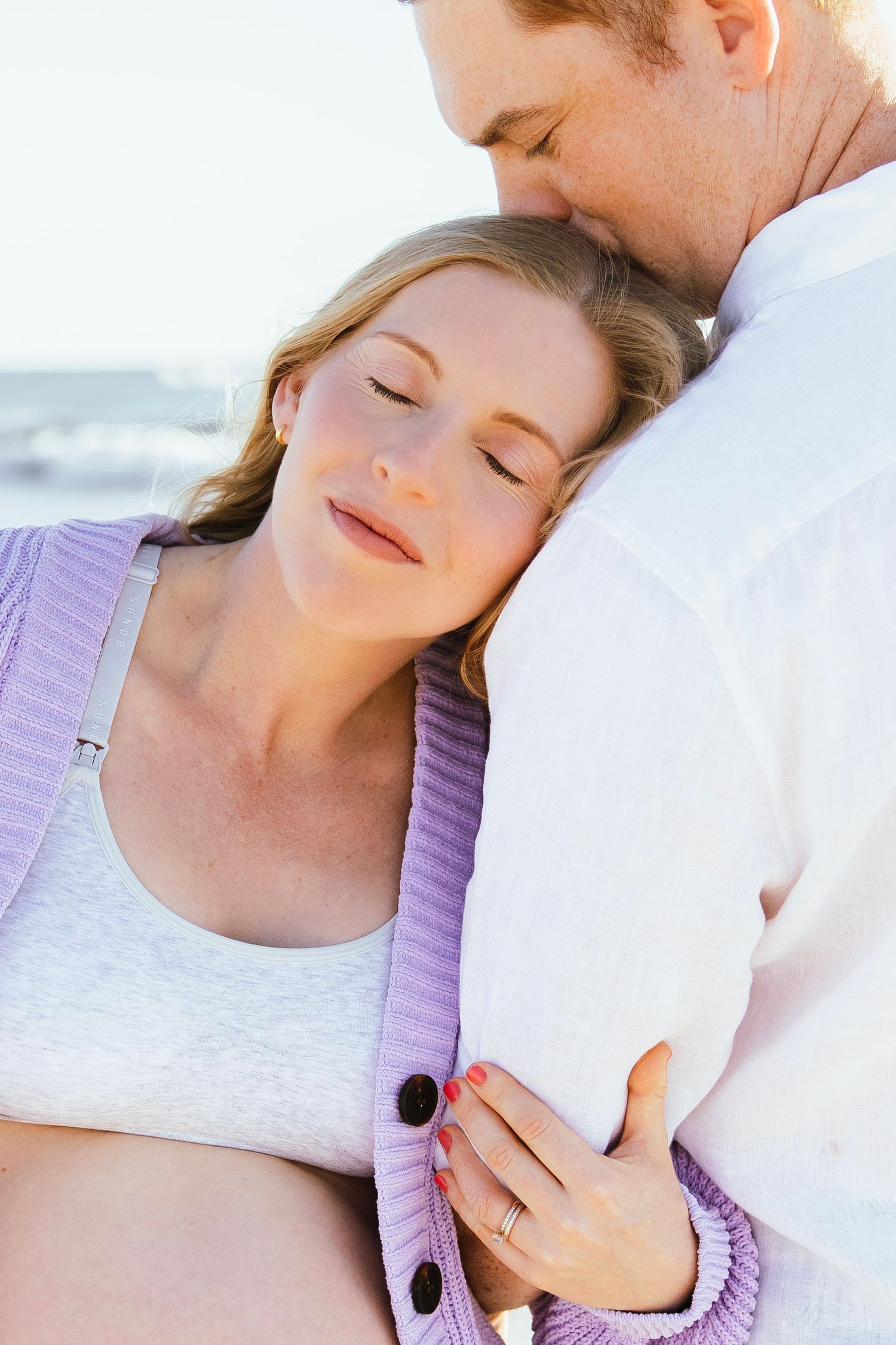 Couple sharing a tender embrace outdoors, woman resting head on man's chest with eyes closed near the ocean.