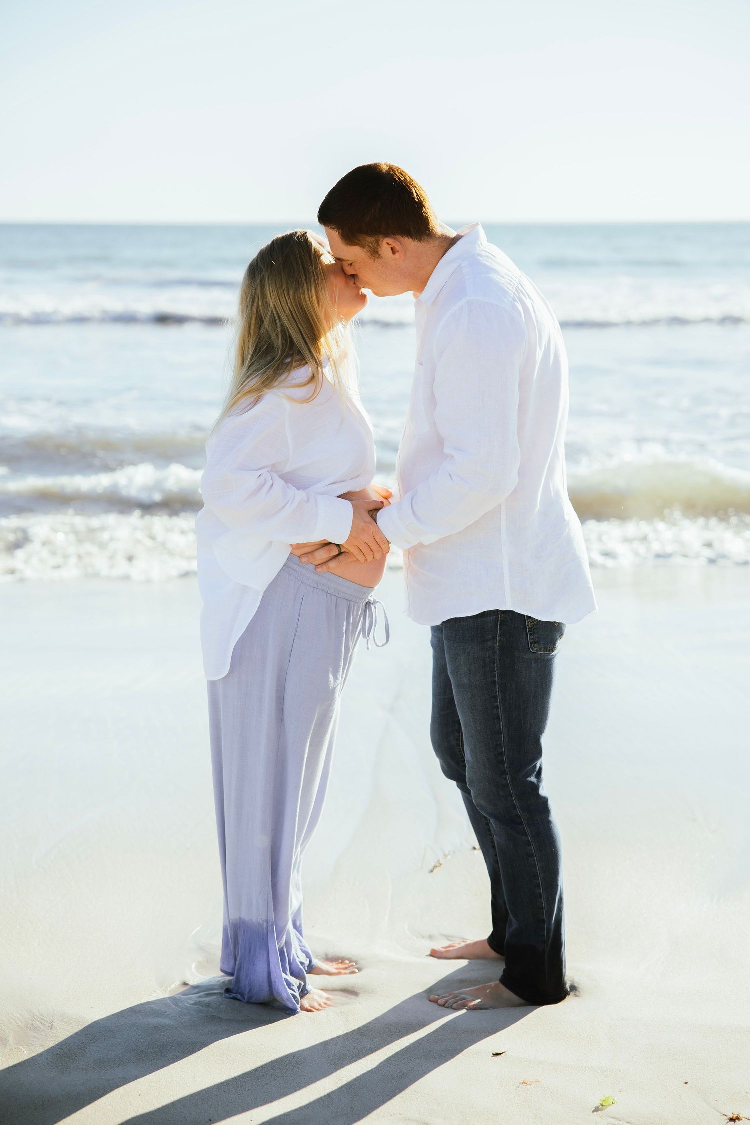 Pregnant couple sharing a kiss on the beach shoreline, waves in the background.