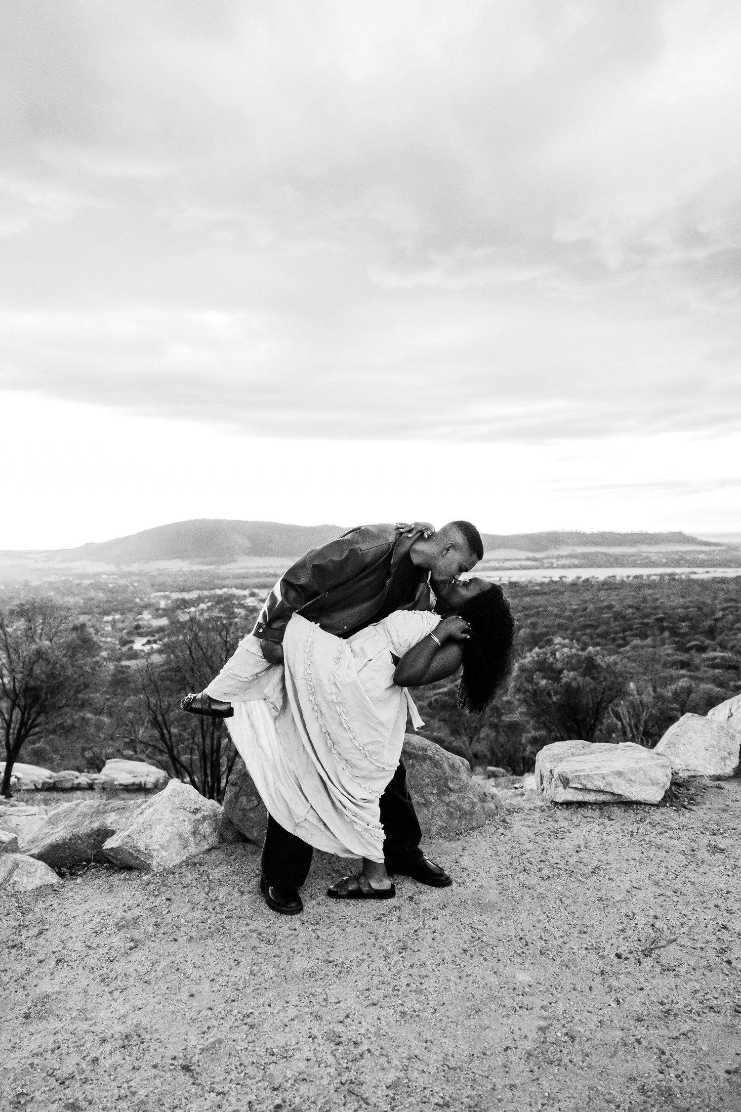 Black and white photo of a couple sharing a passionate dip kiss on a rocky overlook with mountain views.