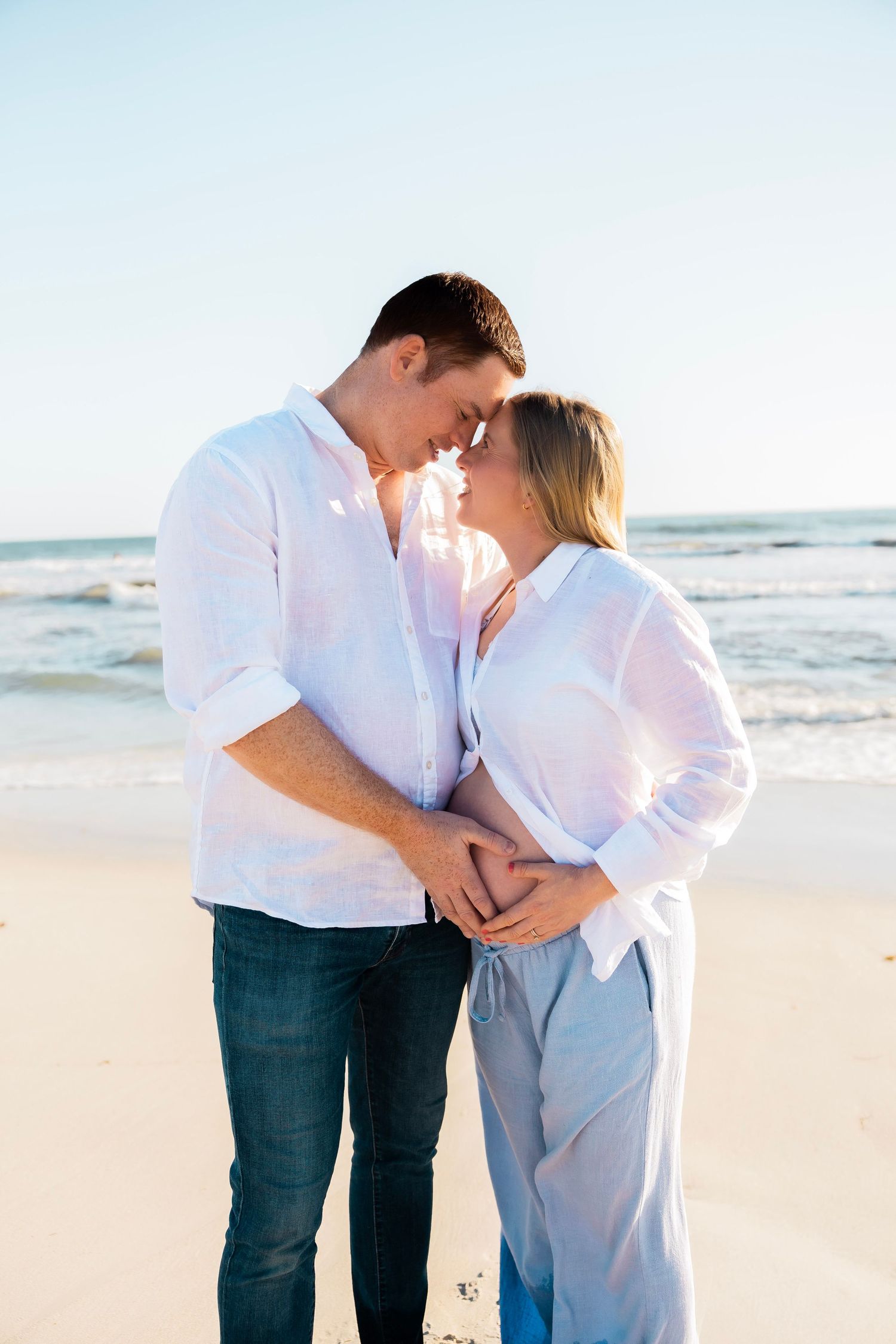 Couple sharing a romantic moment on the beach, touching foreheads and holding pregnant belly near ocean waves.
