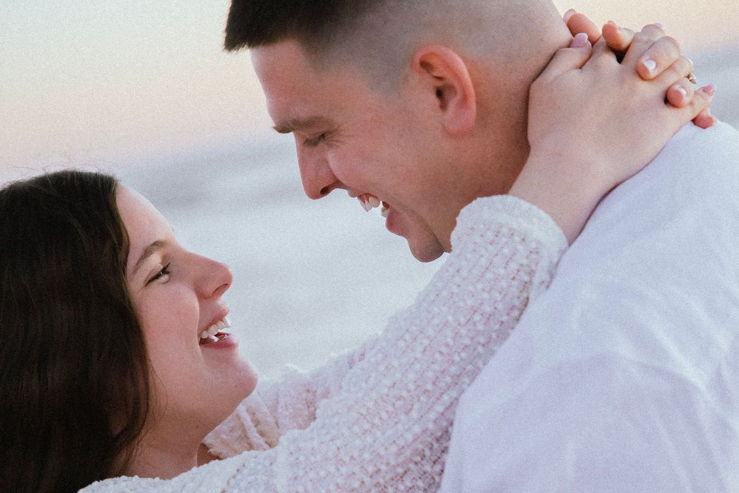 A joyful couple embraces and laughs together on a beach at sunset, the woman wearing a lace top and the man in white.
