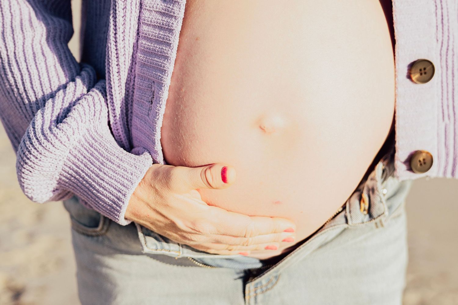 Close-up of a pregnant woman cradling her bare baby bump, wearing a purple cardigan and denim jeans outdoors.