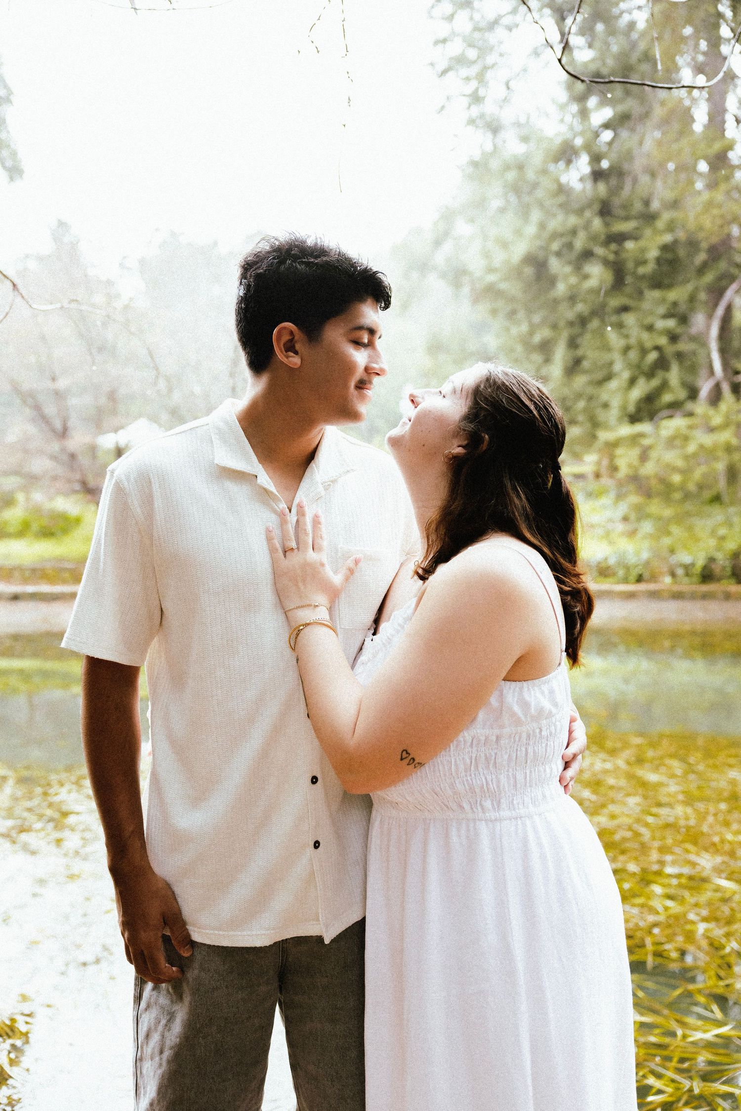 Couple in white outfits share a tender moment by a serene pond in a lush green park.