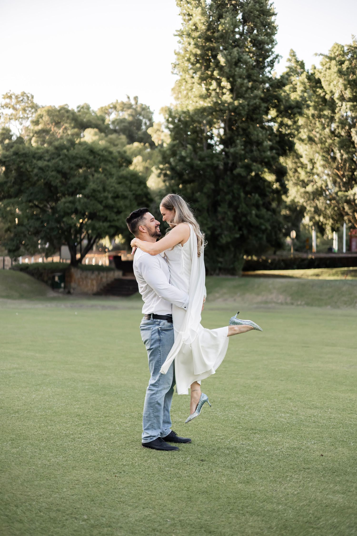 Man lifting woman in white dress as they share a romantic moment on a lush green lawn.