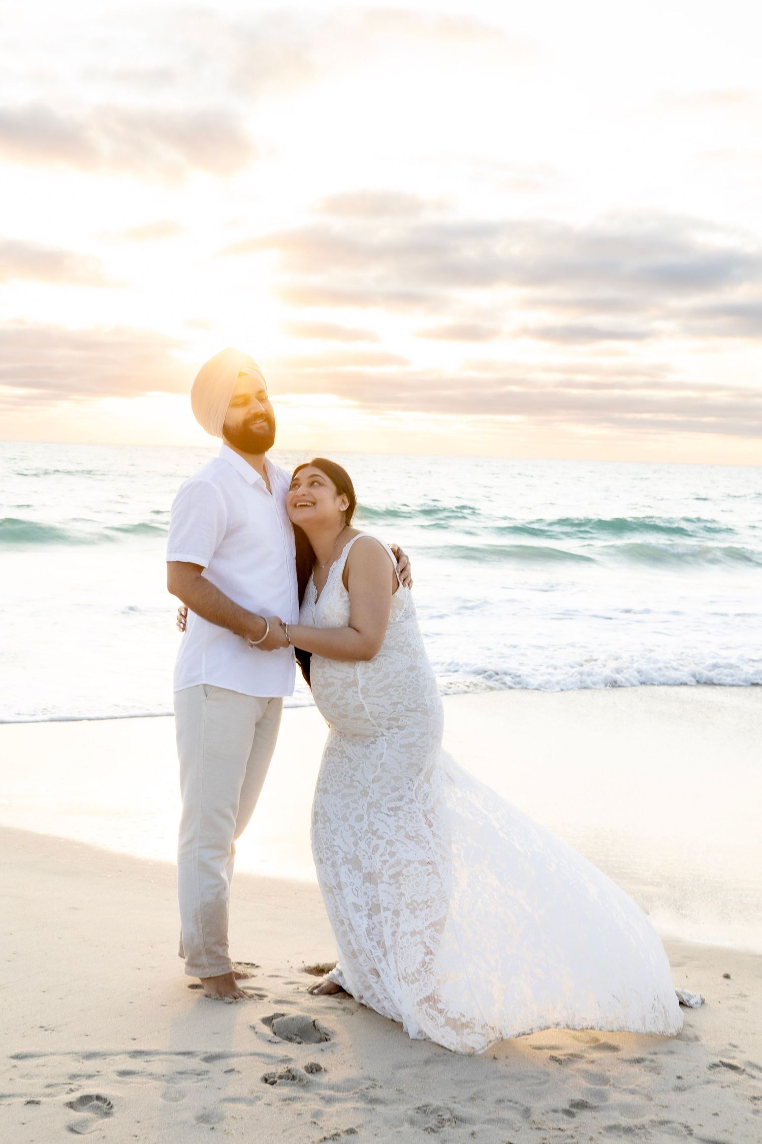 Pregnant couple embracing on sandy beach at sunset, woman in white lace maternity gown looking up lovingly.