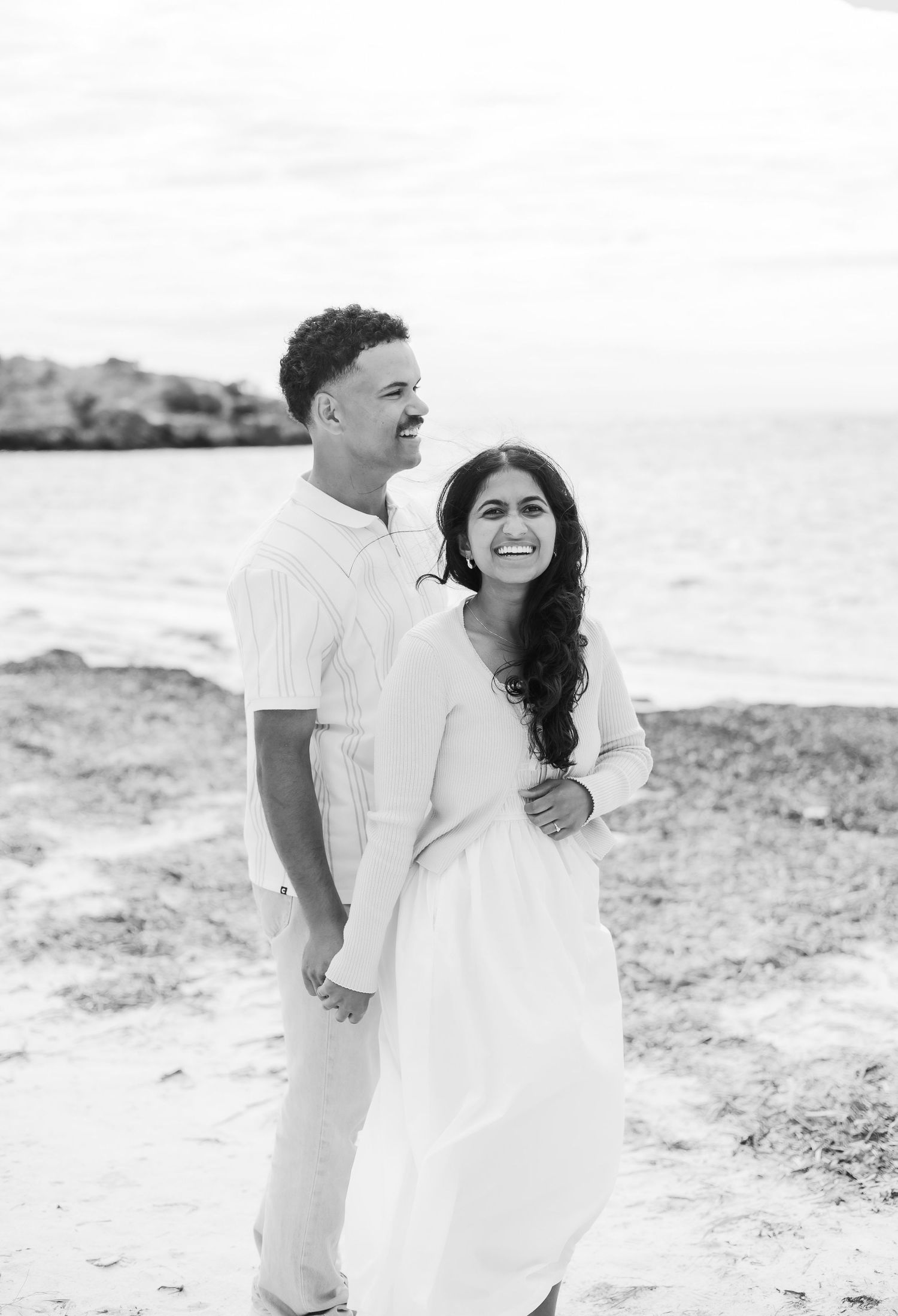 Black and white portrait of a happy couple embracing and smiling together on a peaceful beach shoreline.