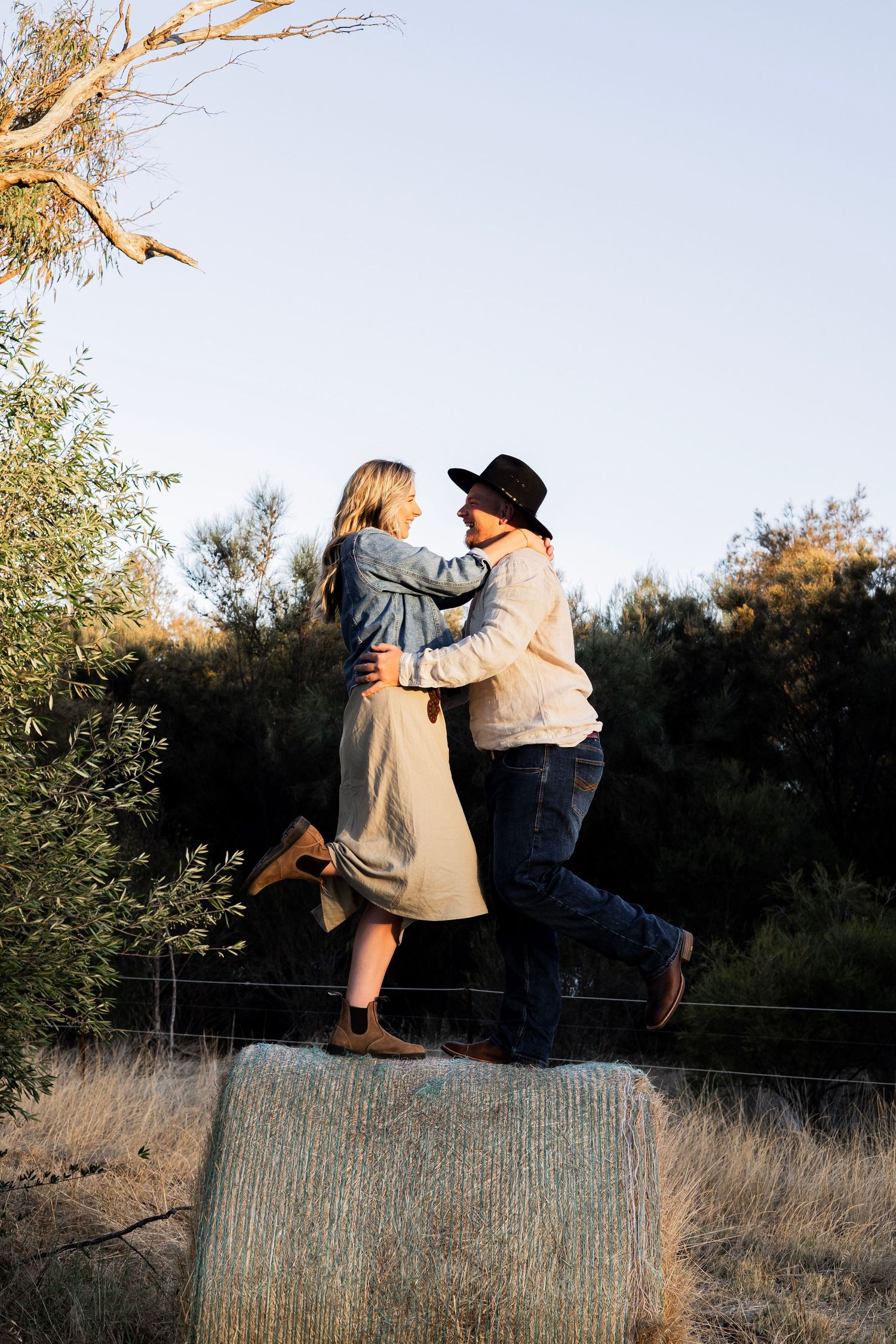 Couple embracing and smiling at each other while standing on a hay bale in a rural setting.