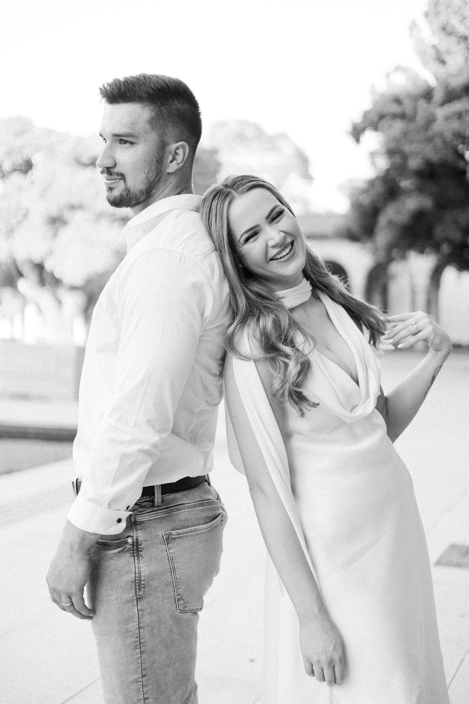 Black and white photo of smiling couple leaning back-to-back outdoors in elegant casual attire.