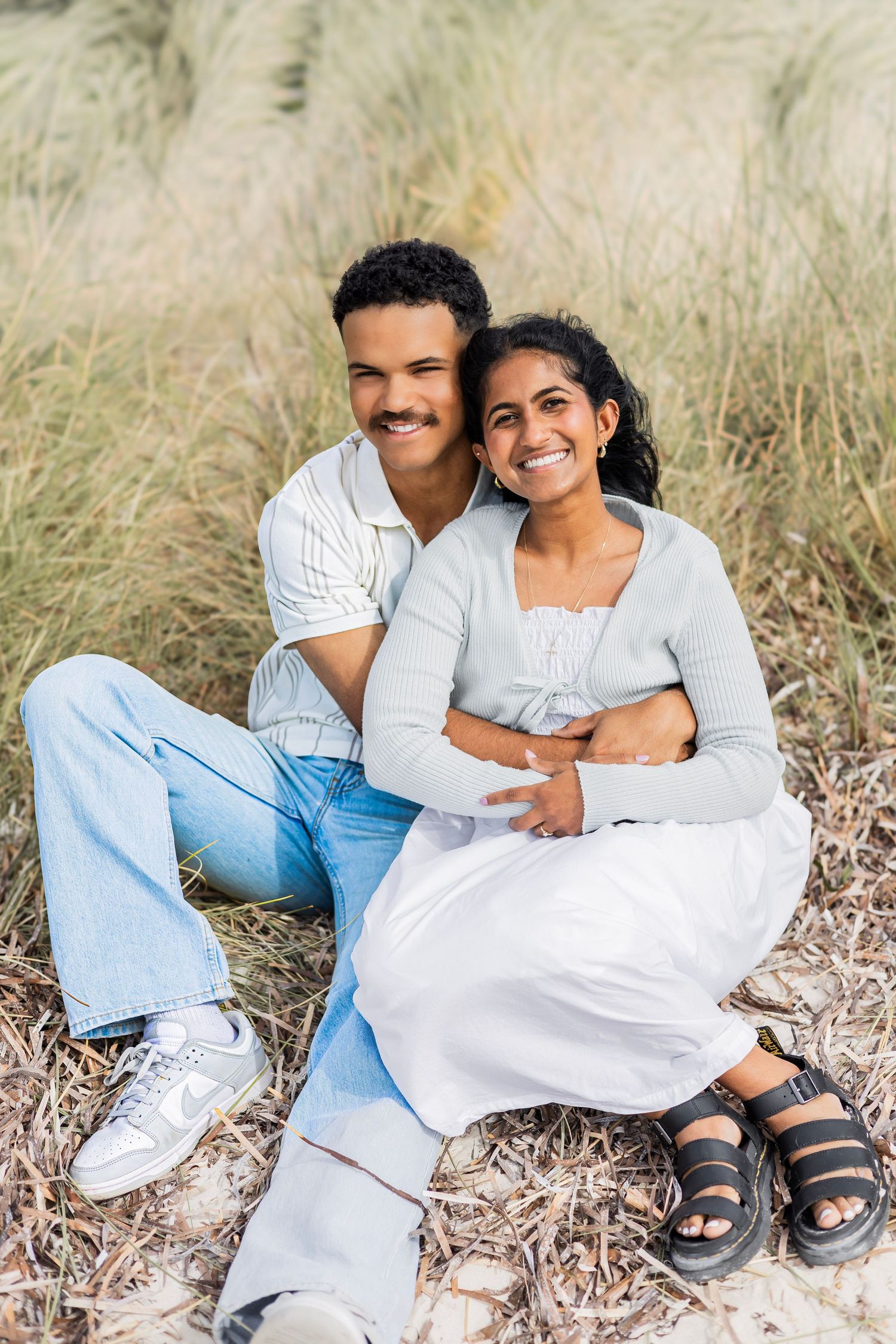 Smiling couple sitting together on sandy beach grass, man holding woman close in casual outfits.