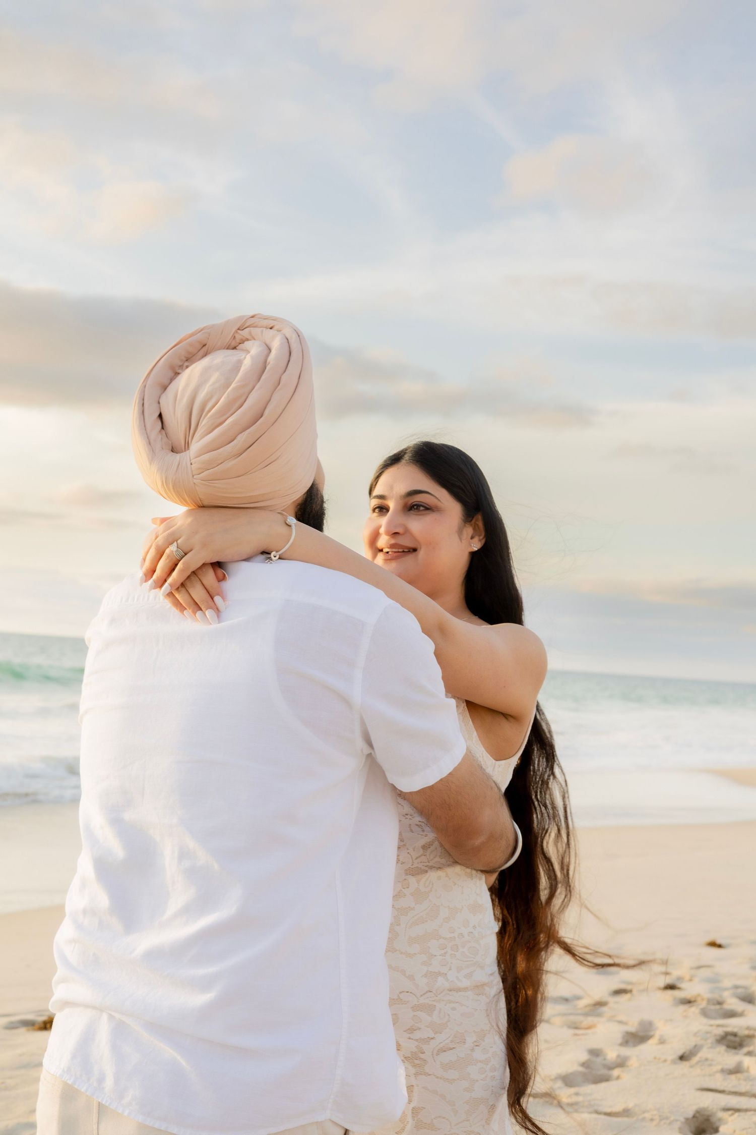 Pregnant woman smiling at her partner on a sandy beach at sunset, both dressed in white.