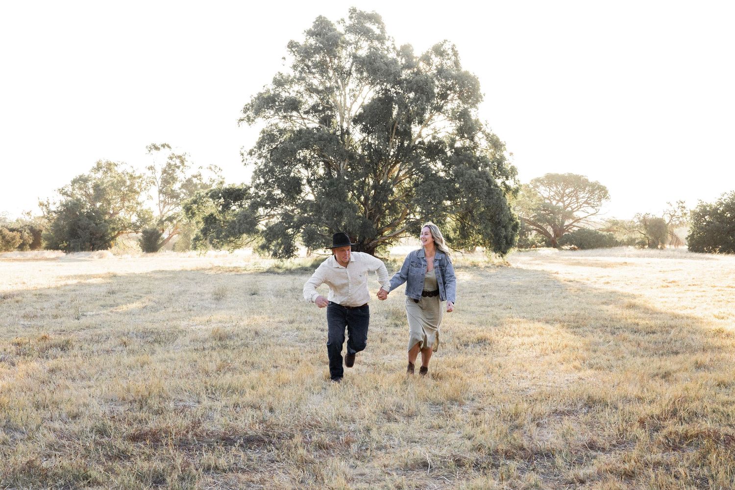 A joyful couple runs hand-in-hand through a golden sunlit field with trees in the background.