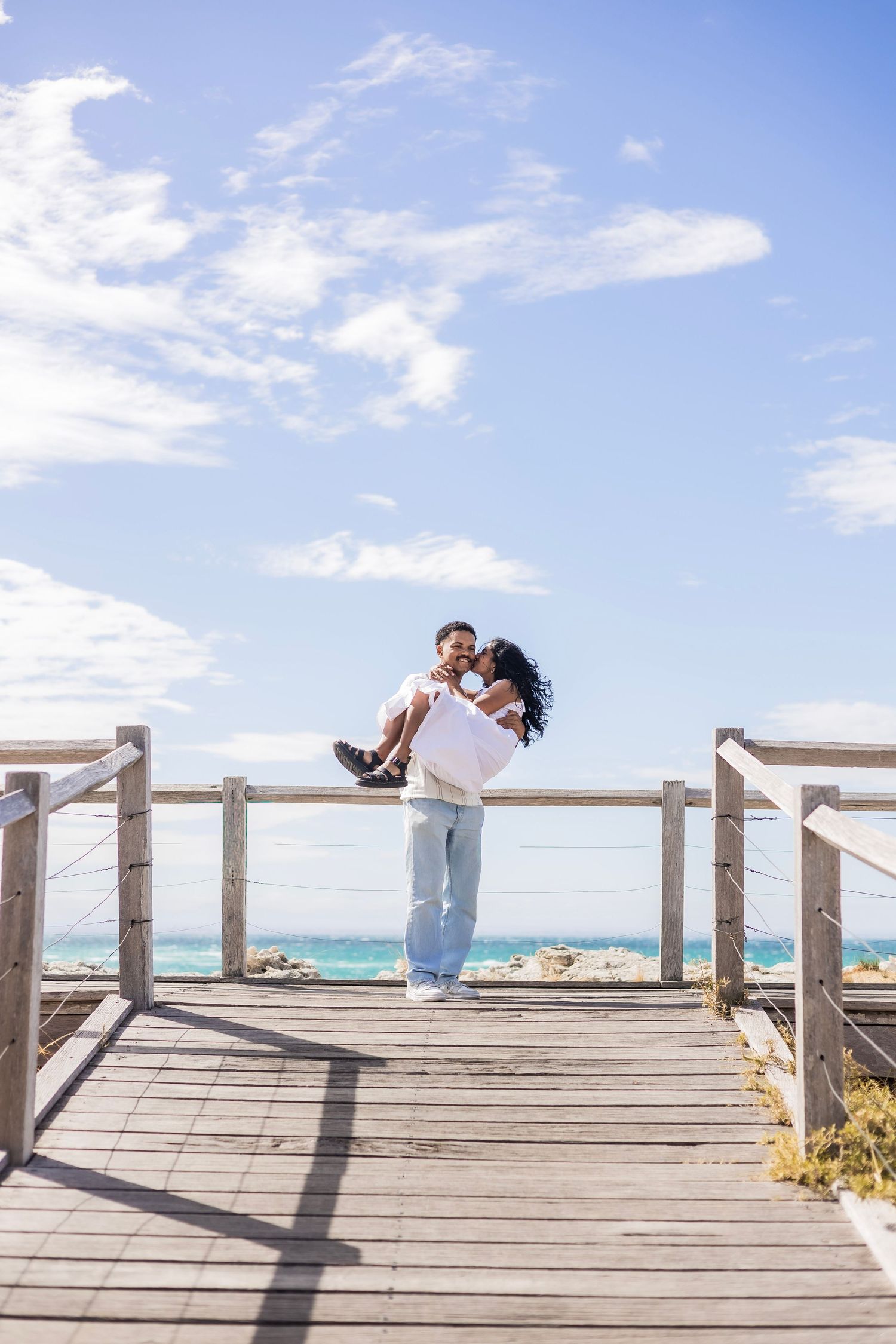 Man carrying woman in his arms on a wooden boardwalk with turquoise ocean in the background.