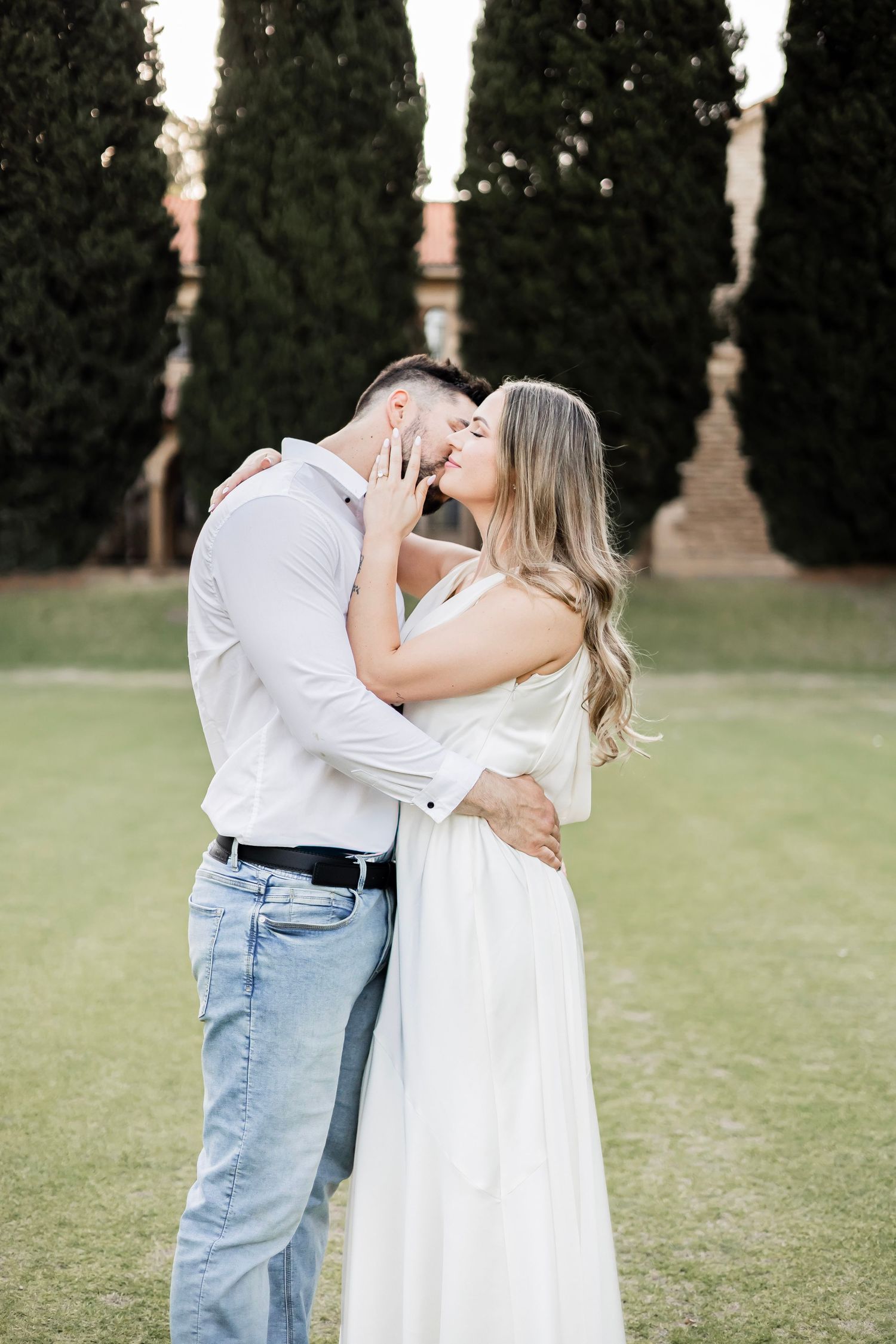Couple sharing a passionate kiss outdoors on green lawn with cypress trees and stone building behind.