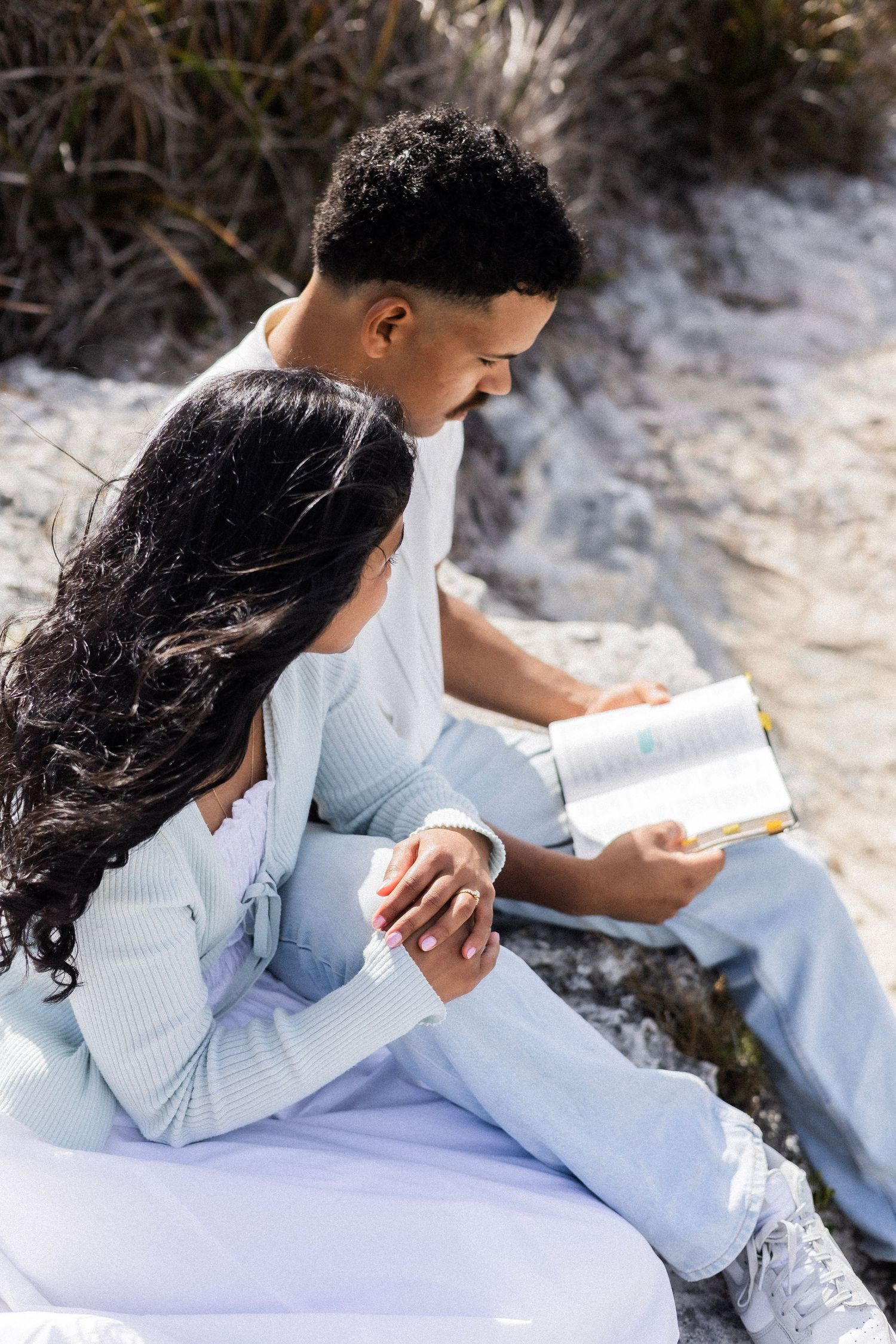 Couple sitting together on sandy rocks reading a Bible, woman leaning against man in pastel outfits.