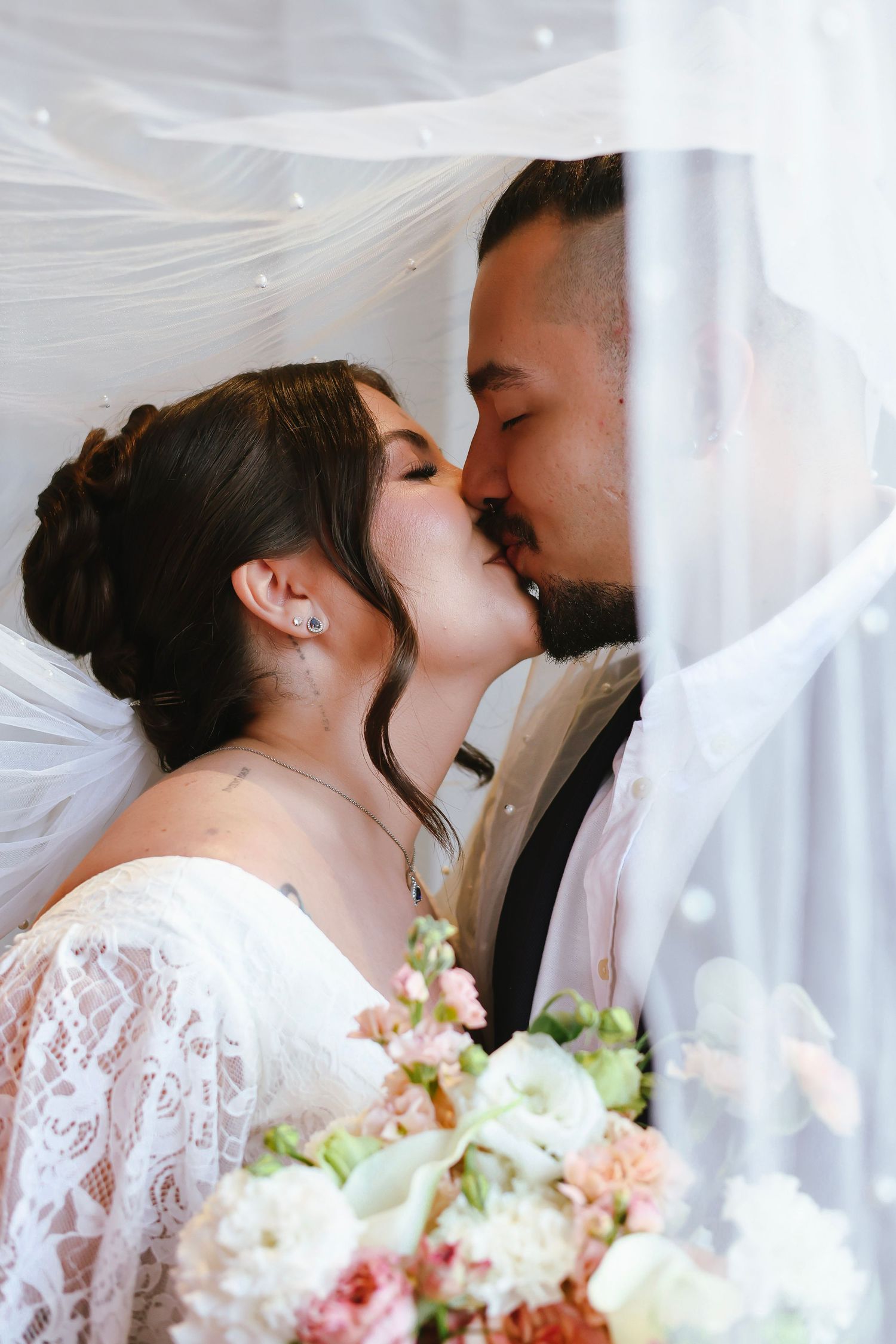 Bride and groom share a romantic kiss under flowing white veil, bride holding pastel floral bouquet.