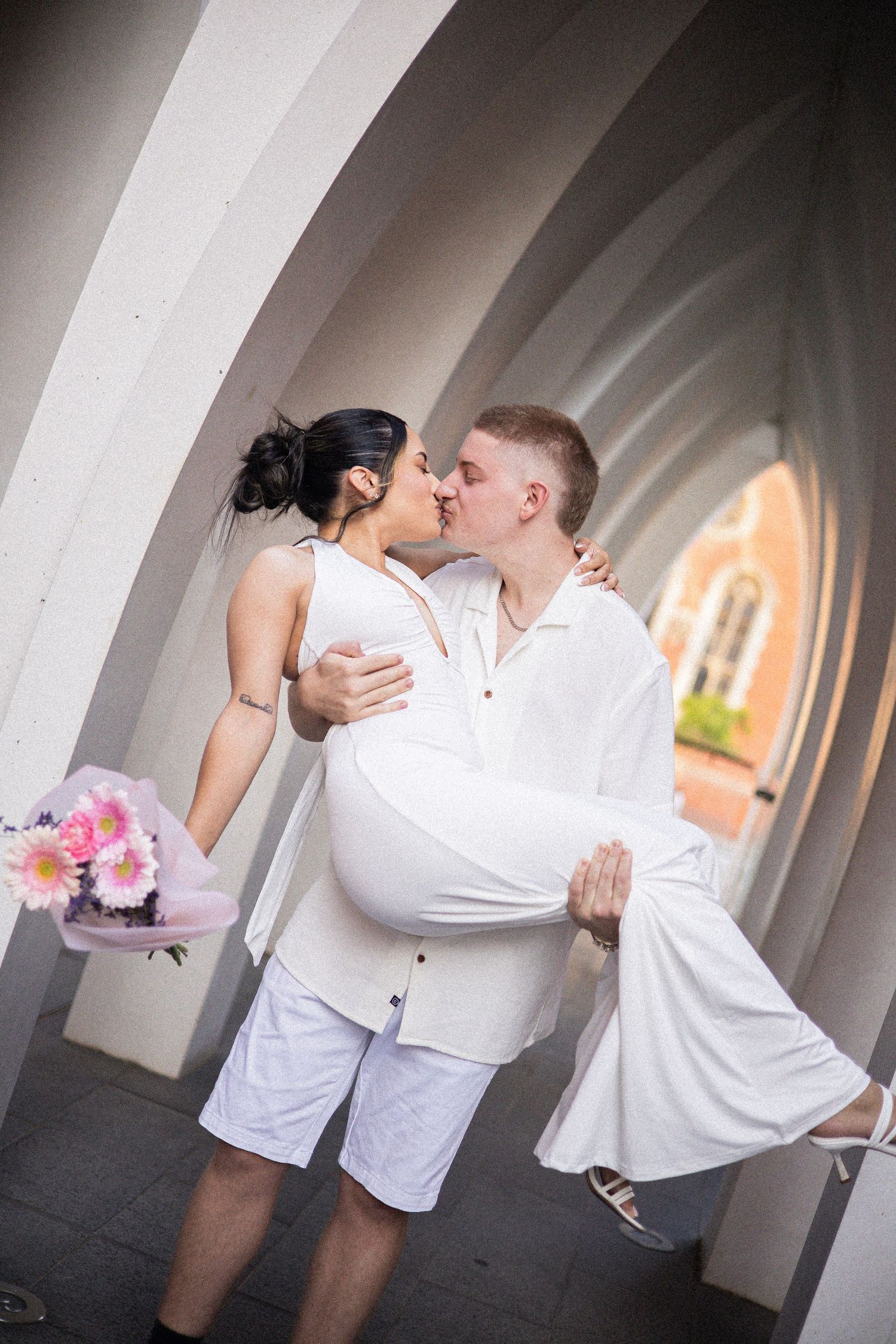 A couple in white attire shares a romantic kiss under arched columns, the woman holding a pink flower bouquet.