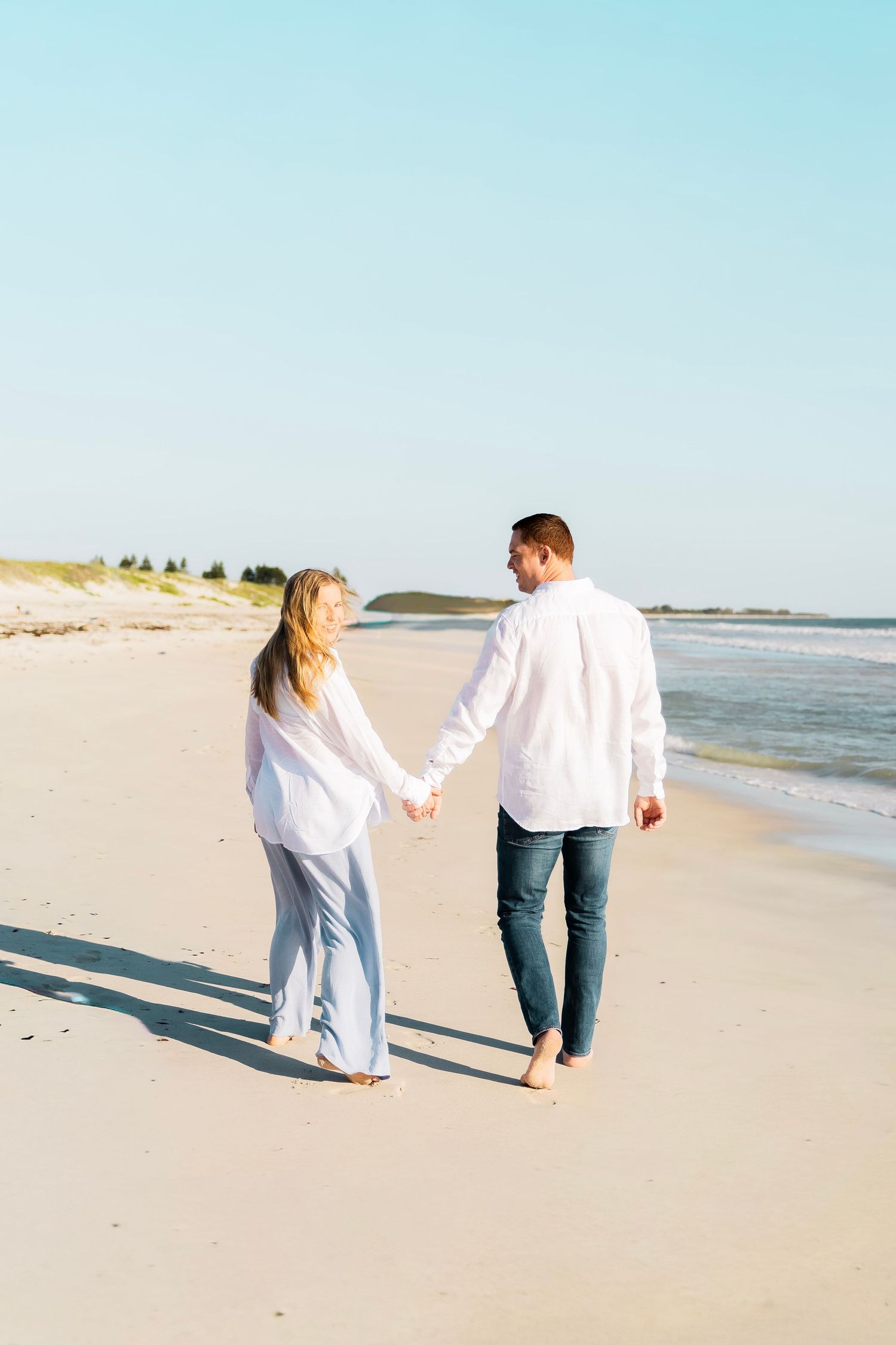 Couple holding hands walking barefoot on a sunny sandy beach, both wearing white shirts, viewed from behind.