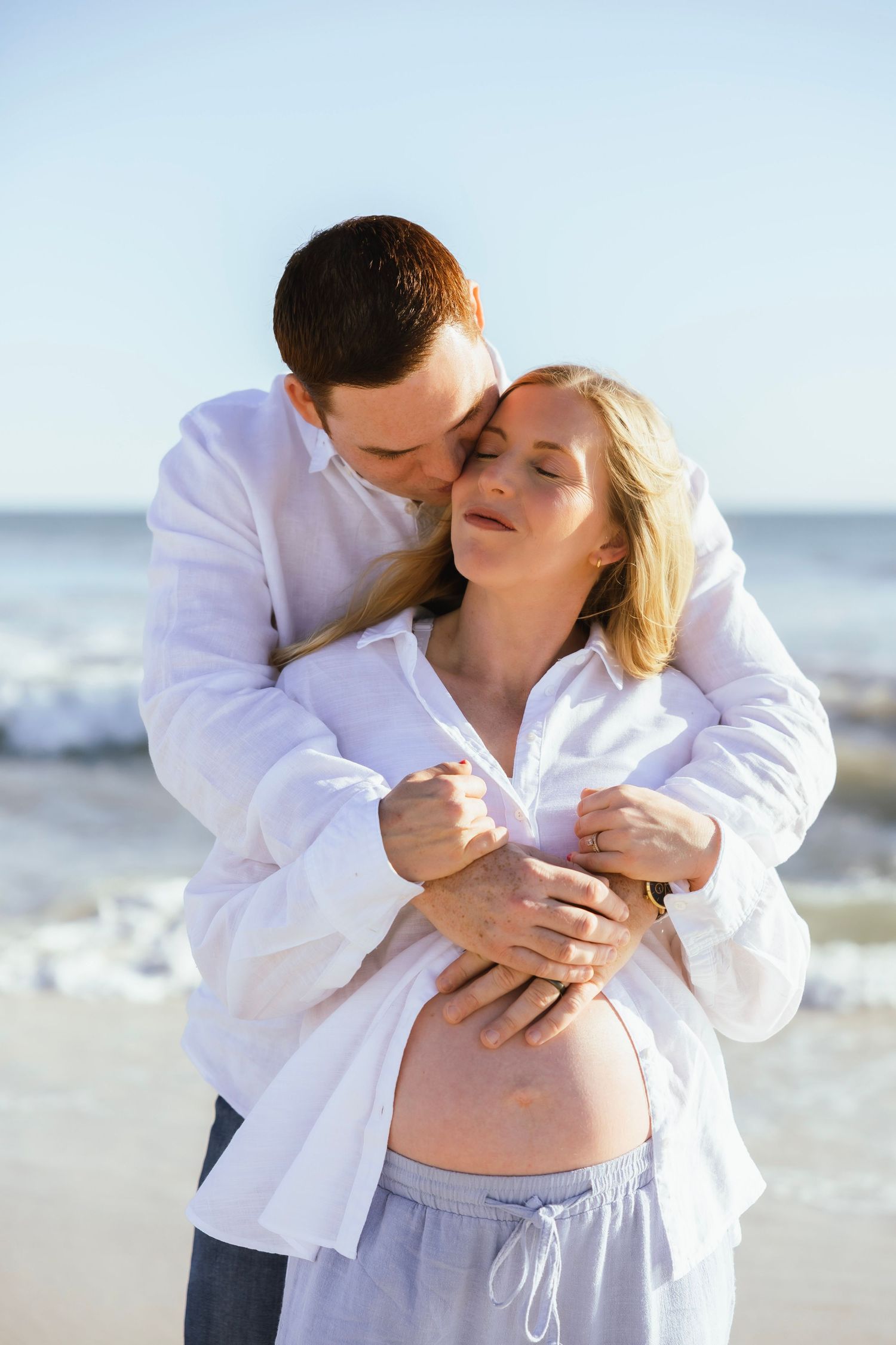 Couple embracing on beach, pregnant woman in white shirt showing baby bump, man kissing her head near ocean.