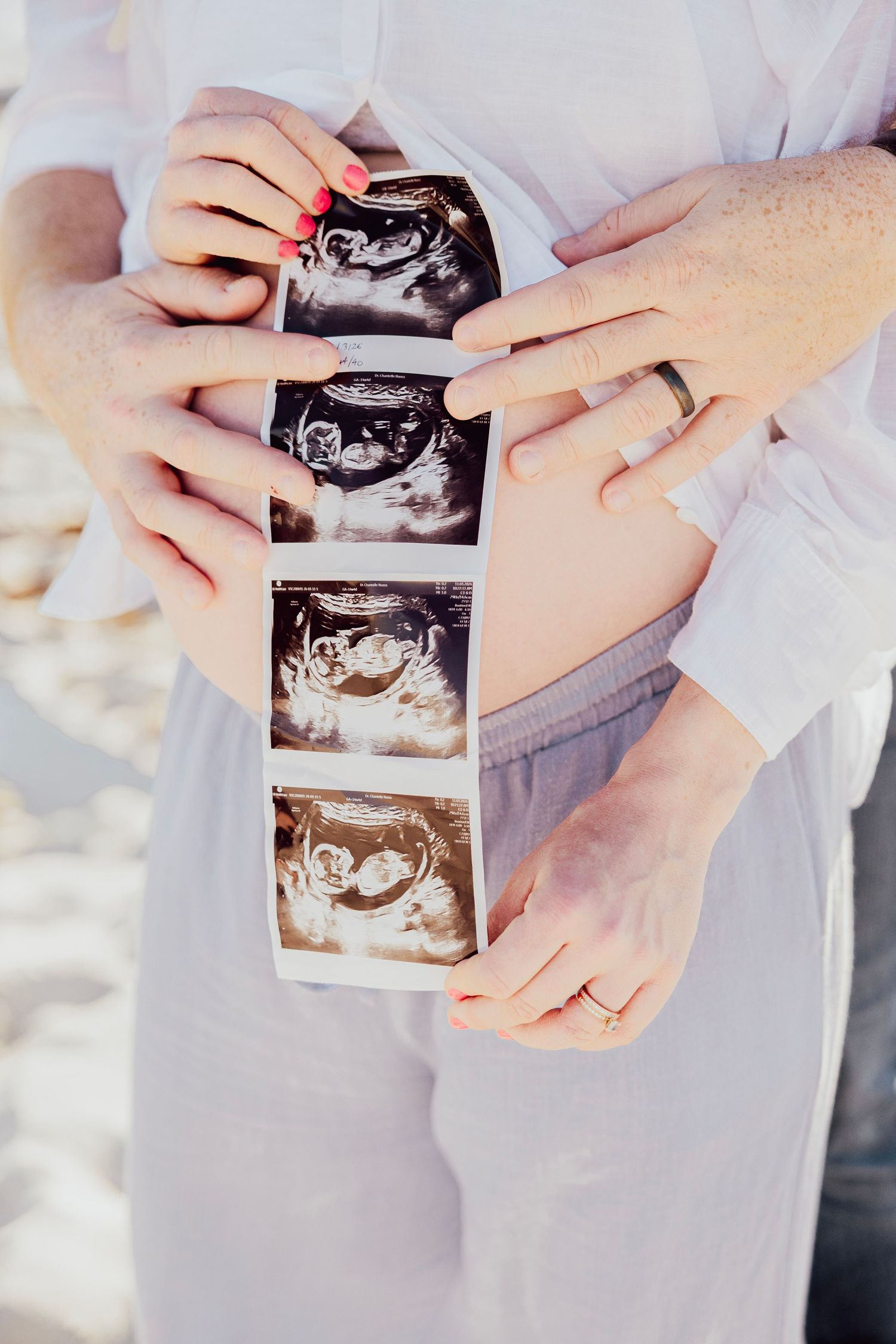 Couple holding ultrasound photos against pregnant belly on beach.