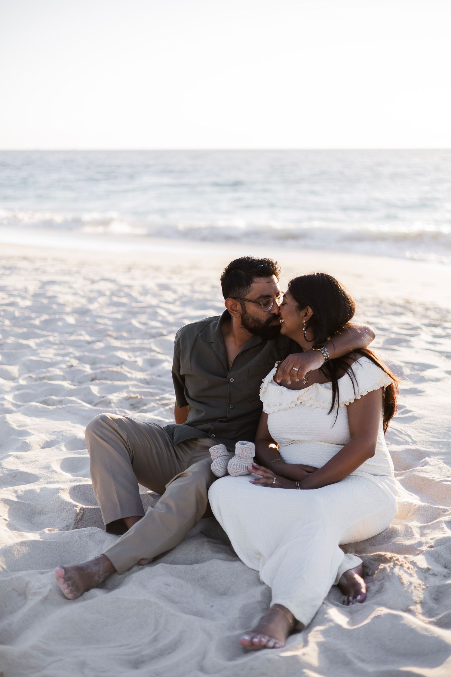 Expecting couple sitting on beach sand holding baby booties, sharing a tender moment at sunset.