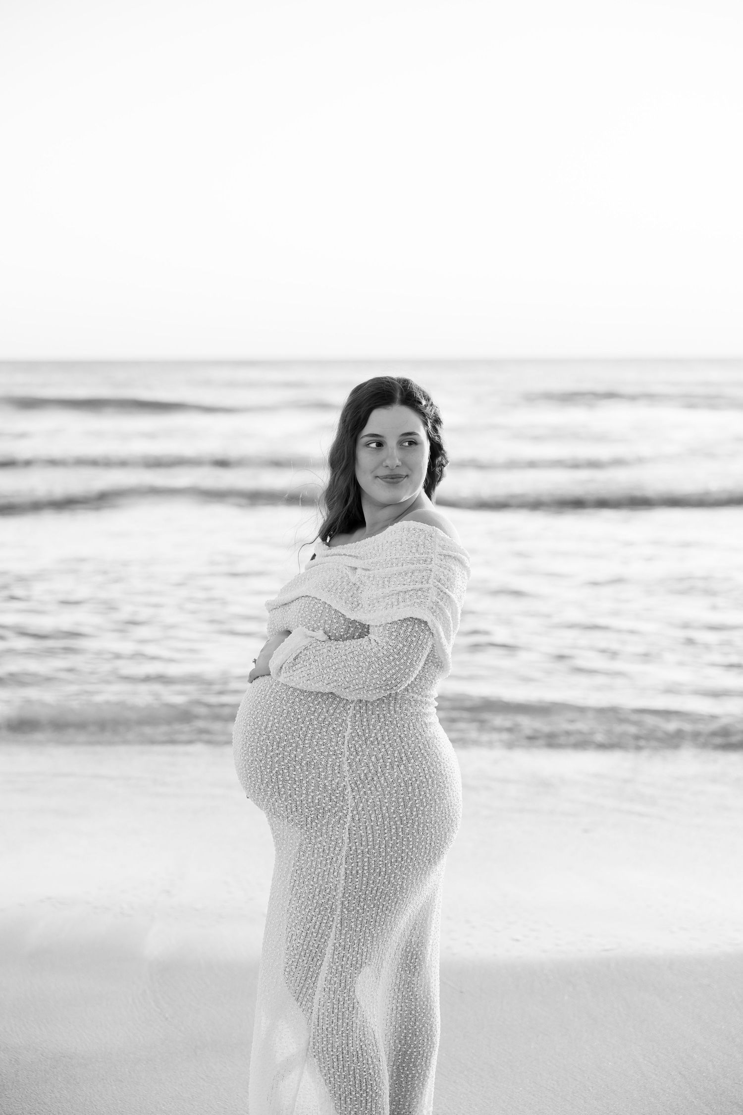 Black and white maternity photo of pregnant woman in lace dress cradling bump on beach.