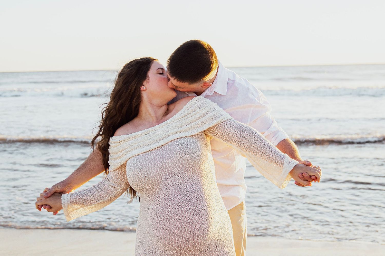 Couple sharing a romantic kiss on the beach, woman in white lace dress with ocean waves in the background.