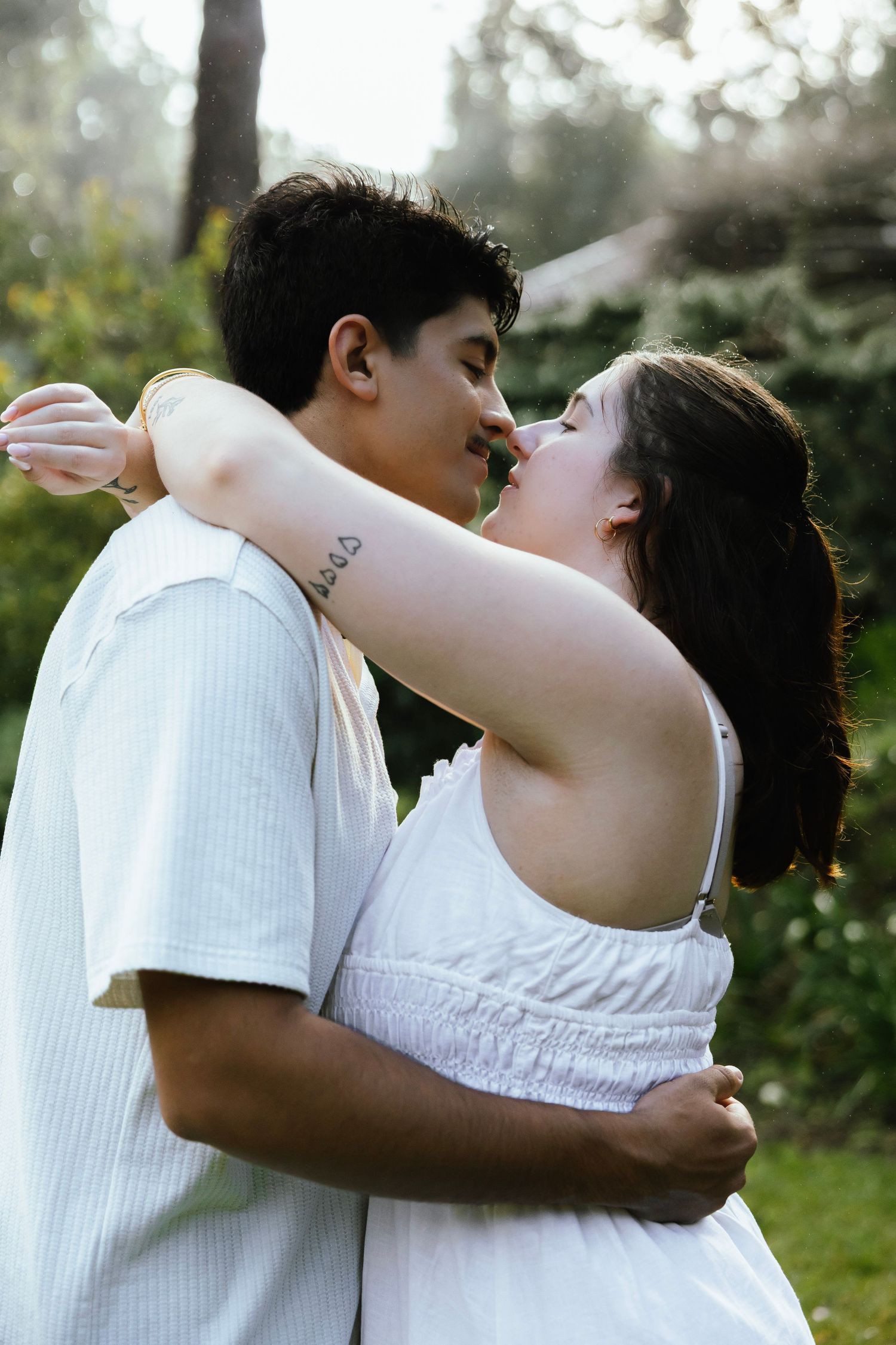 Young couple embracing and about to kiss outdoors, woman in white dress with arm tattoo visible.