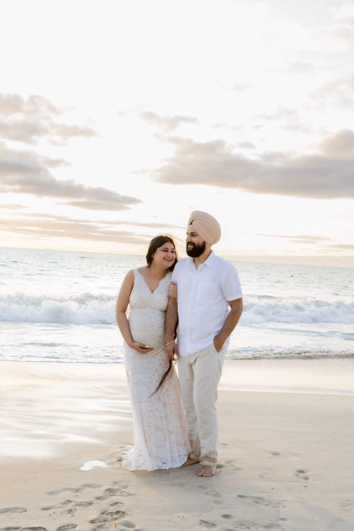 Expecting couple in white outfits sharing a tender moment on sandy beach at sunset.