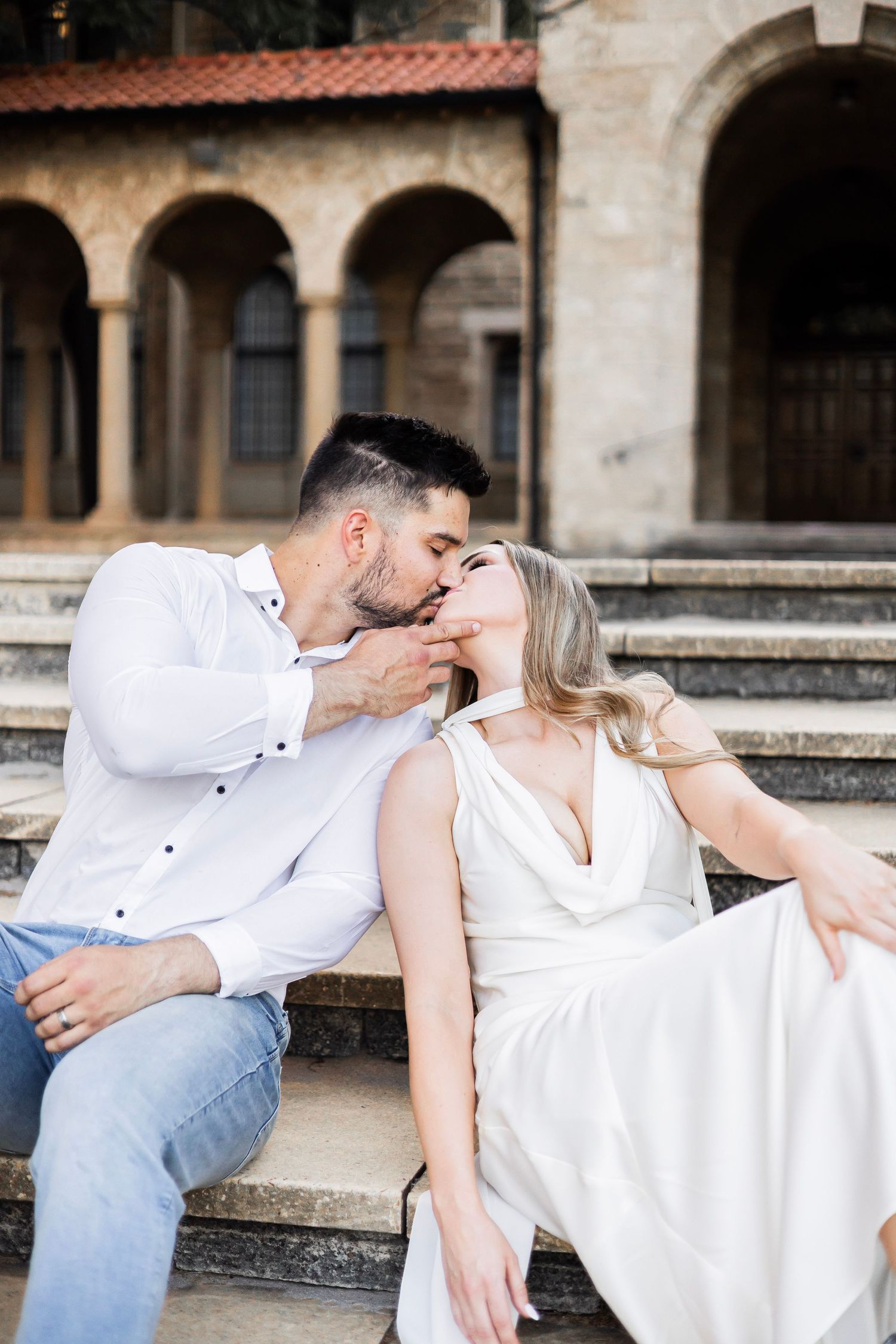 Couple sharing a passionate kiss while seated on stone steps outside a historic building with arched colonnade.