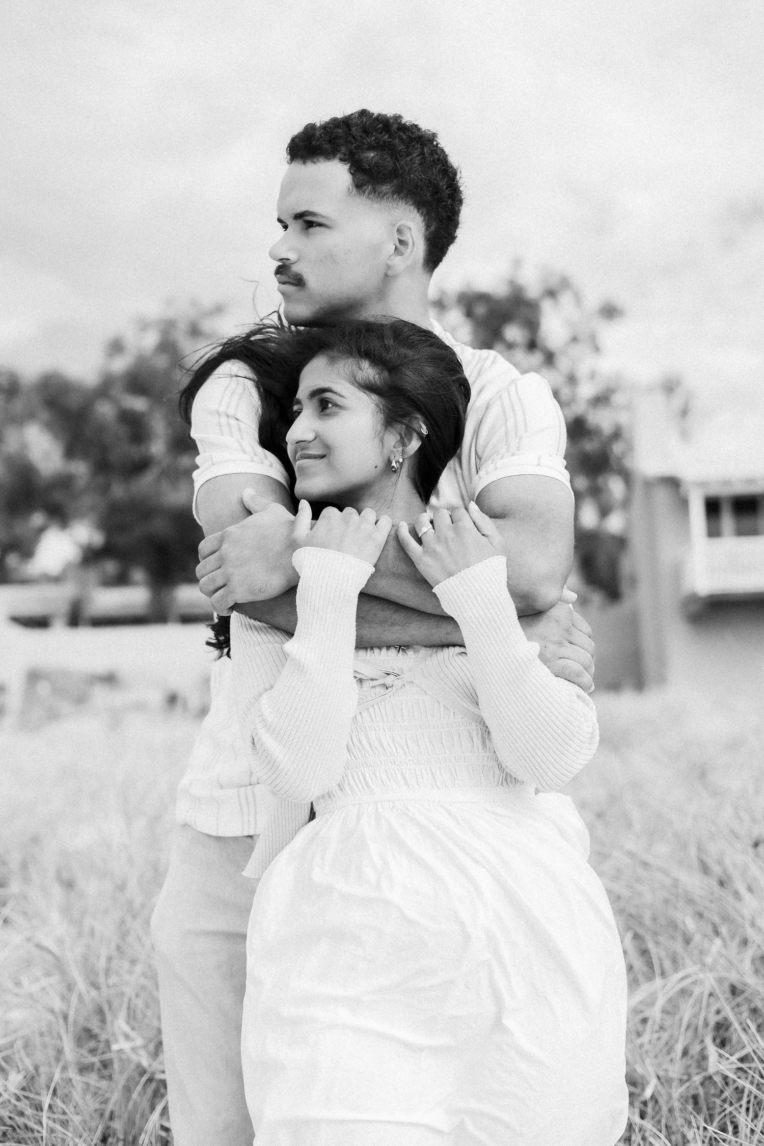 Black and white portrait of couple embracing in field, man holding woman from behind tenderly.