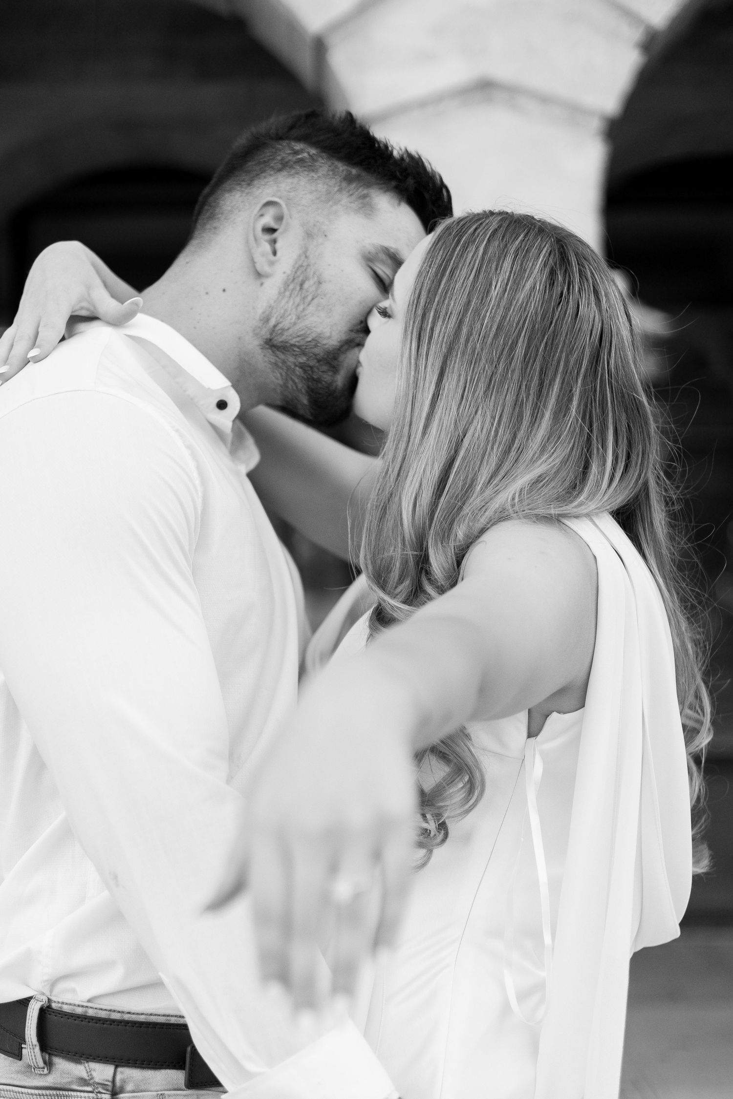 Black and white romantic close-up of couple sharing a kiss under arched stone architecture.