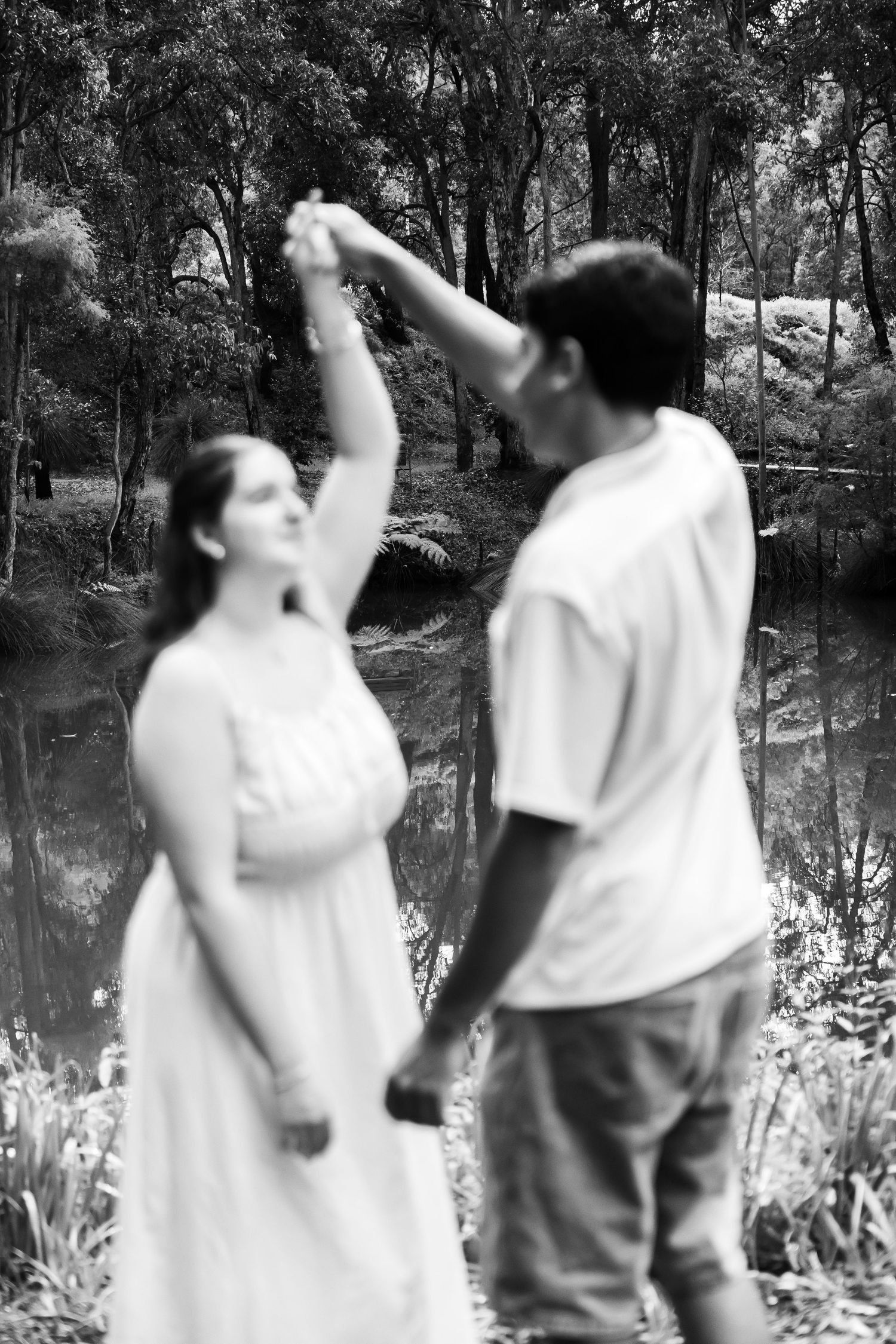 A couple dances outdoors near a reflective pond surrounded by lush trees in a dreamy black and white portrait.