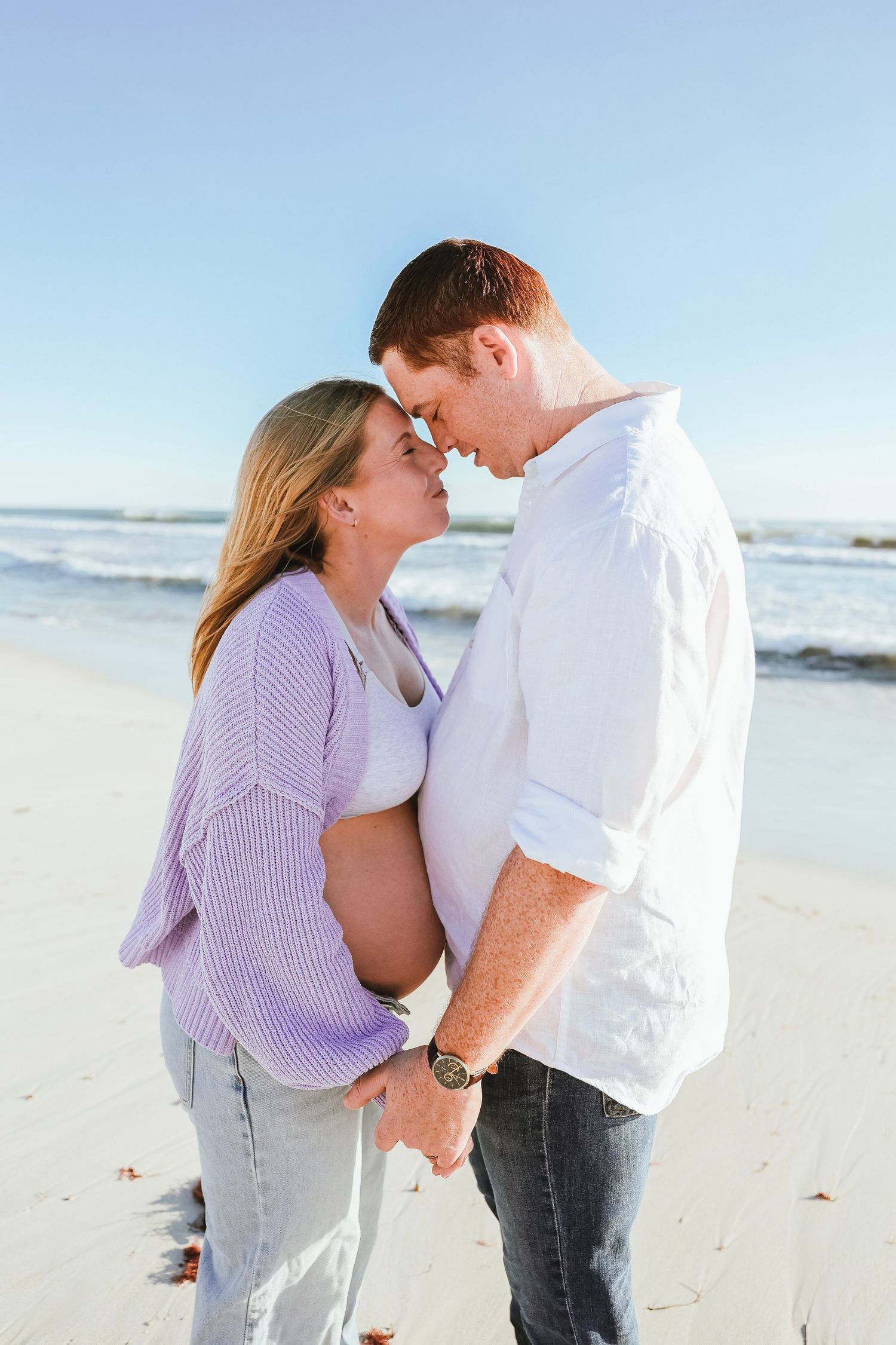 Pregnant woman in lavender cardigan and partner in white shirt touching foreheads tenderly on a beach.