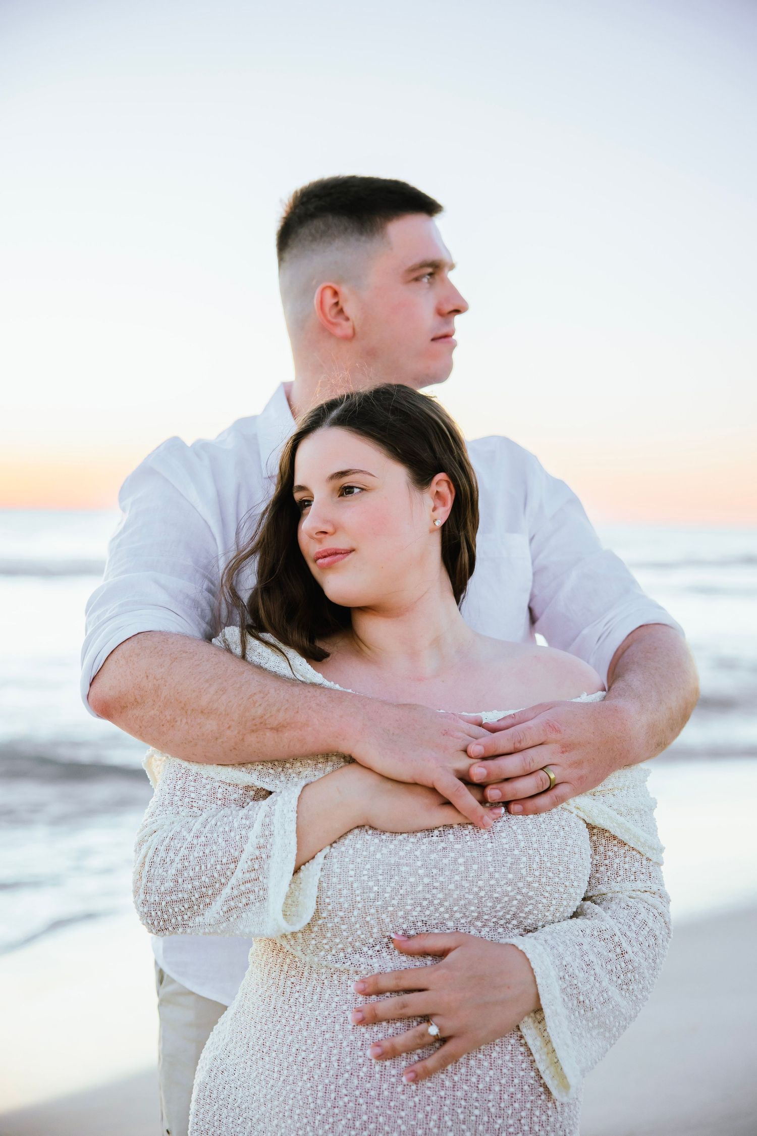Couple embracing on beach at sunset, man in white shirt holding pregnant woman from behind.
