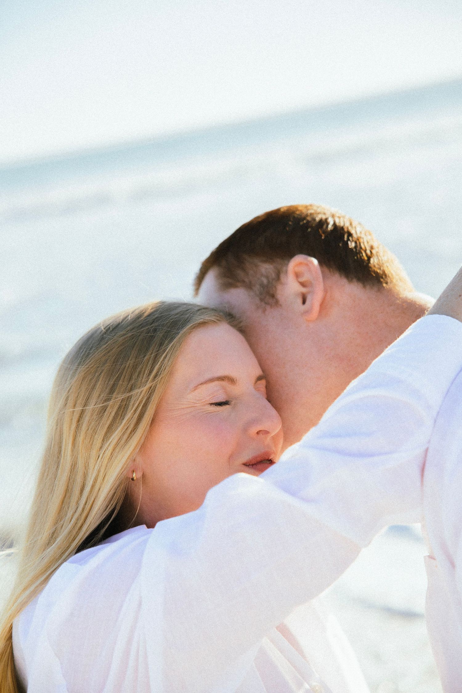 Romantic couple in white shirts sharing a tender moment on the beach, woman with blonde hair closing her eyes.