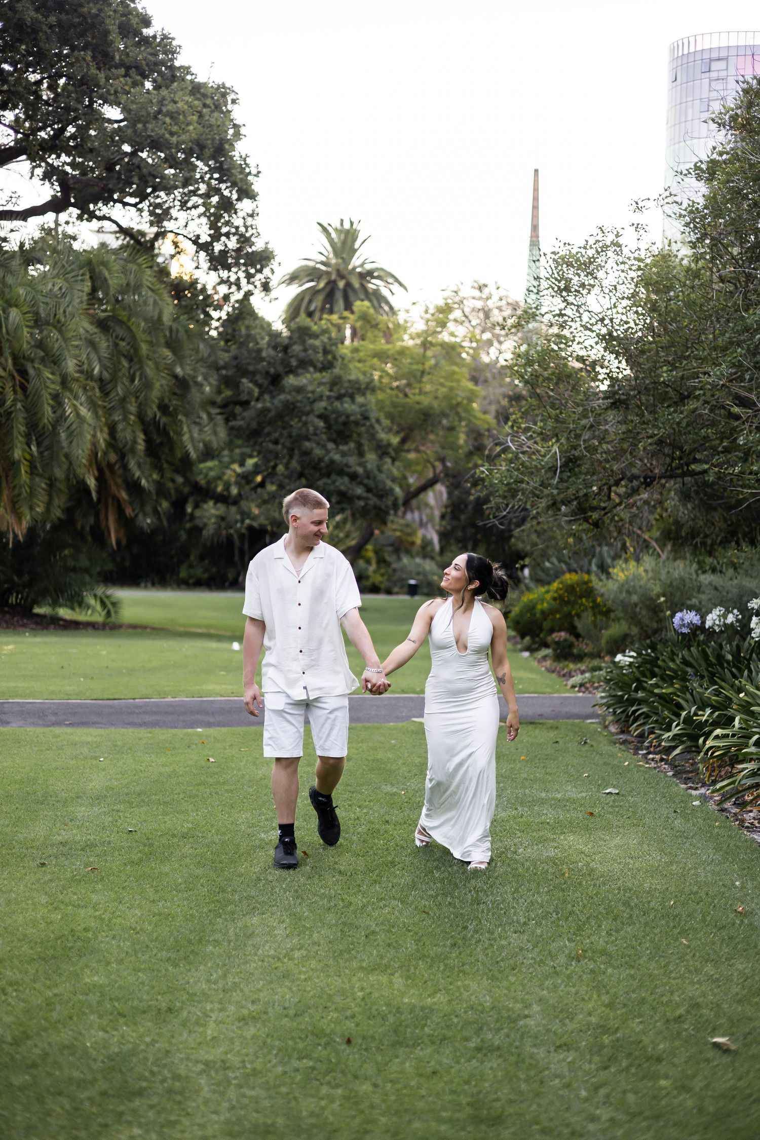 Couple in white outfits holding hands walking through lush green park gardens.