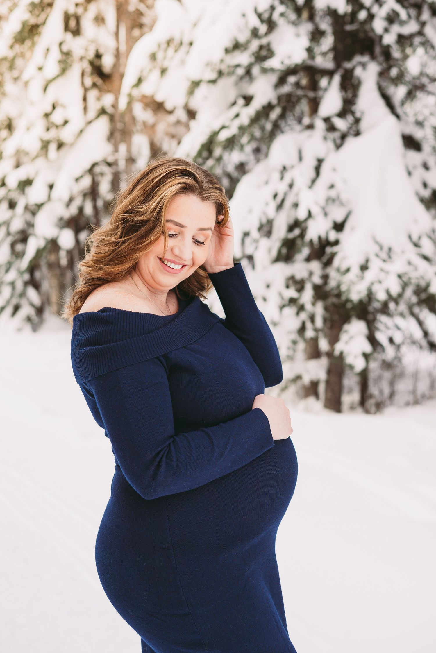 Pregnant woman wearing a dark blue dress standing outdoors in a snowy winter setting during a maternity session