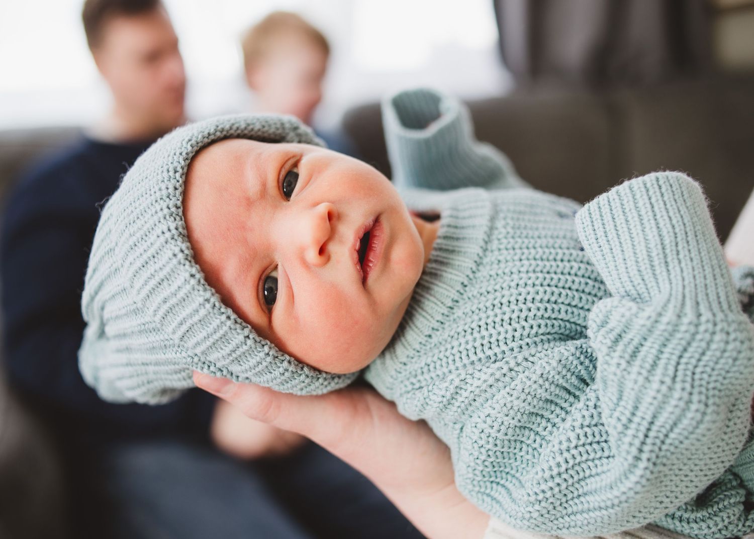 Awake newborn baby wearing a blue knit hat and sweater while being held by a parent